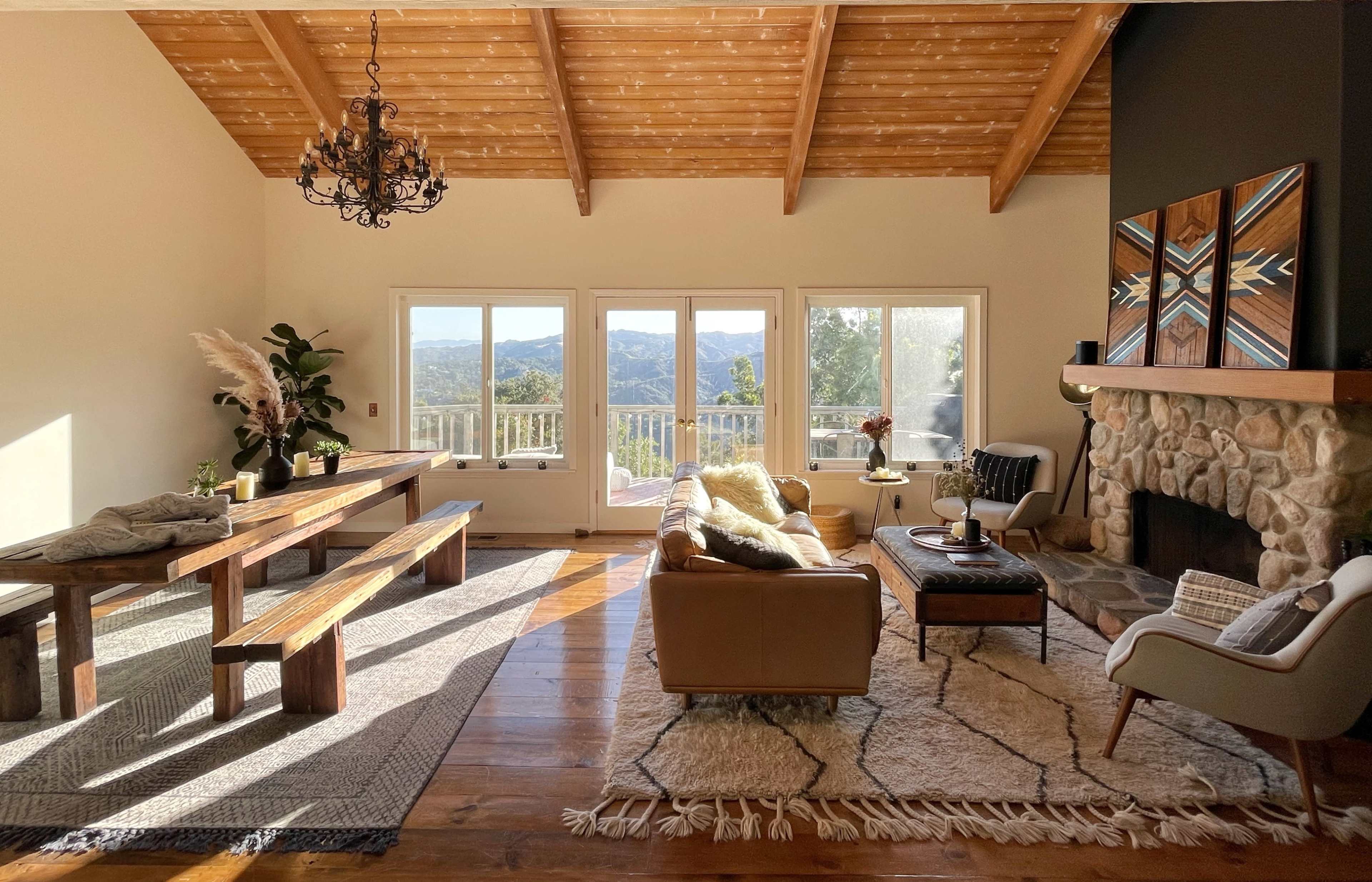 A spacious living room features a wooden dining table, a leather couch, and a stone fireplace with large windows overlooking a scenic view.