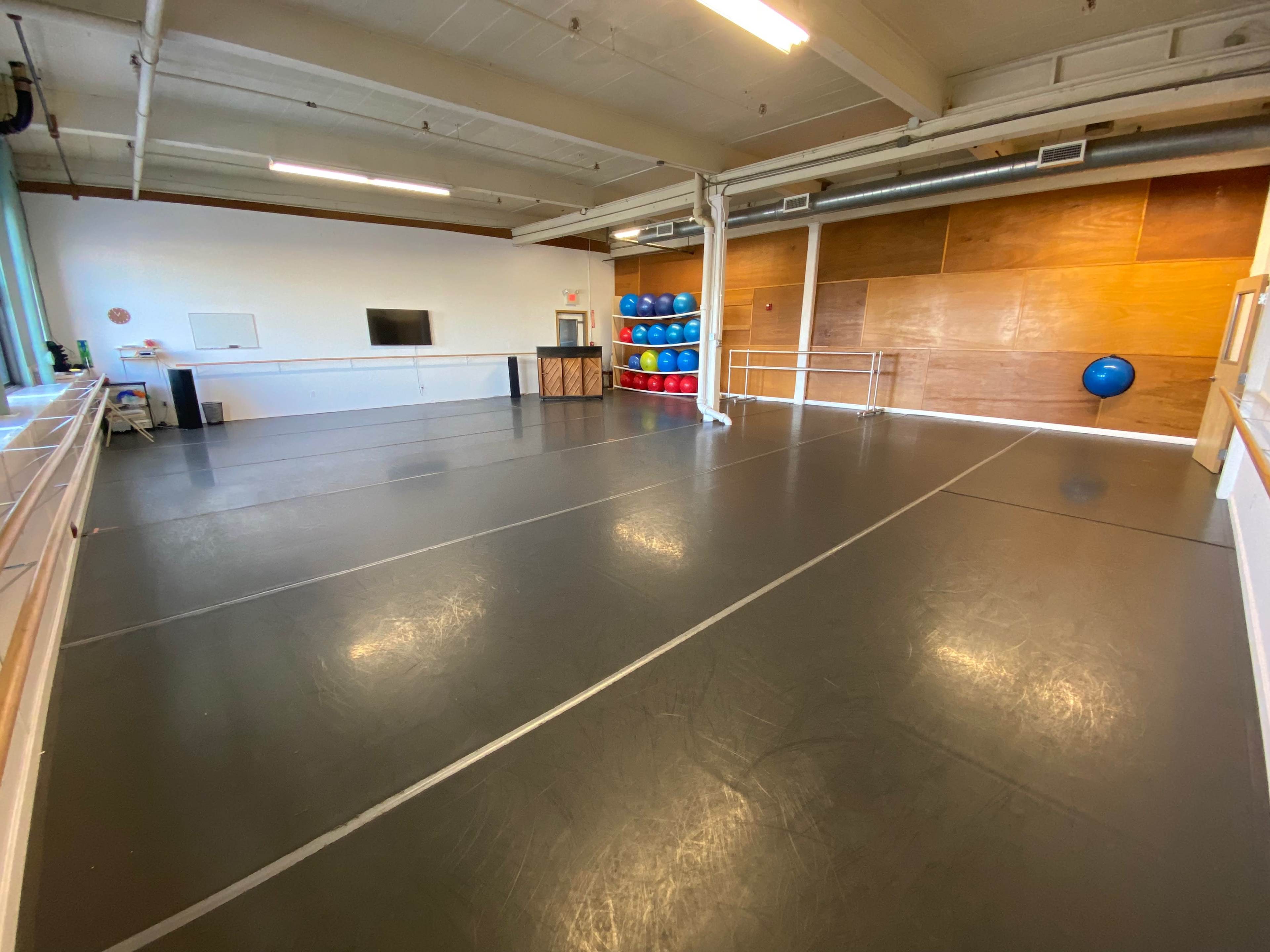 The image shows a spacious fitness studio with a smooth gray floor, mirrored walls, and various fitness equipment stacked in the corner.