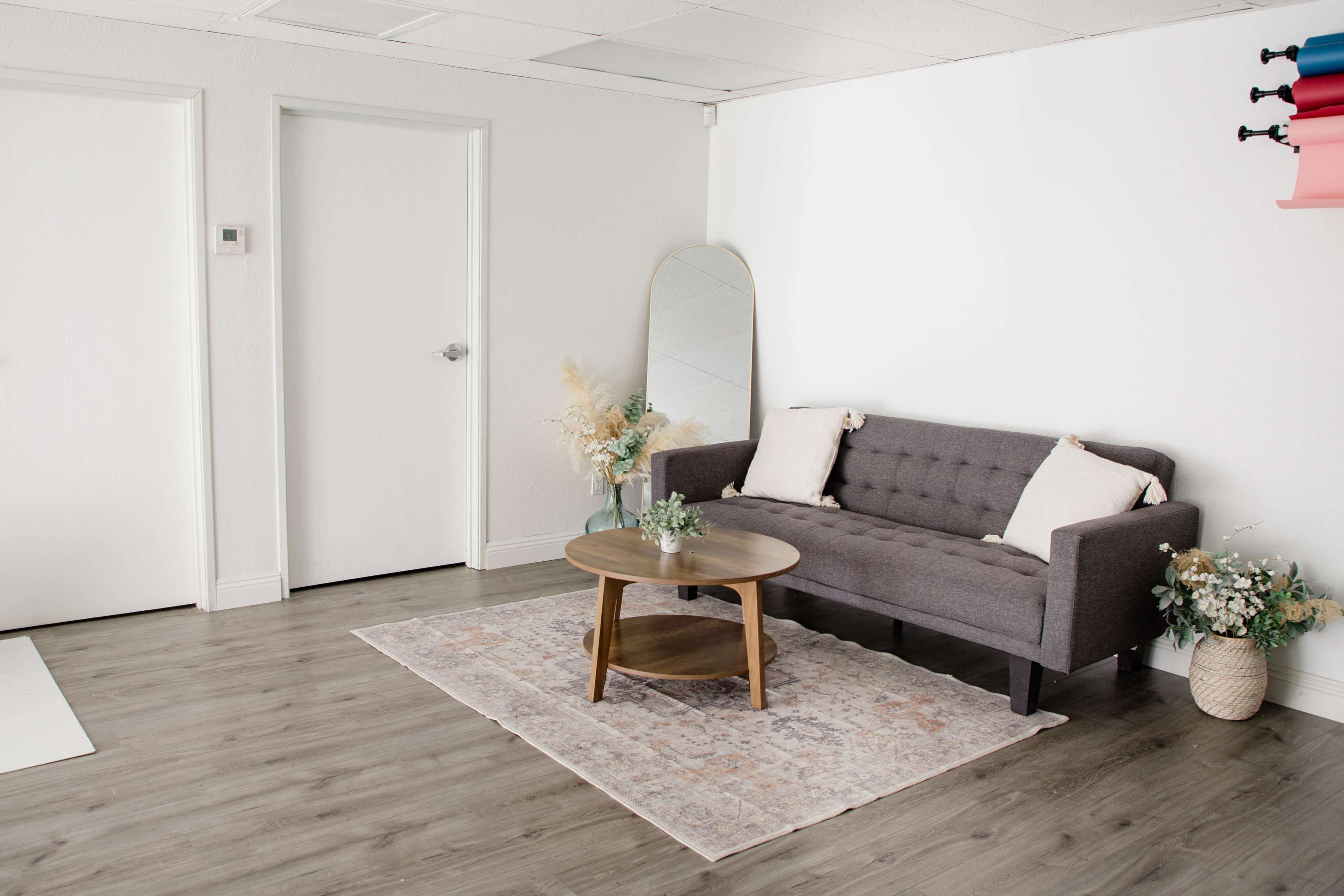 The image shows a modern living room with a gray sofa, a round wooden coffee table, a decorative rug, and a large mirror against a white wall.