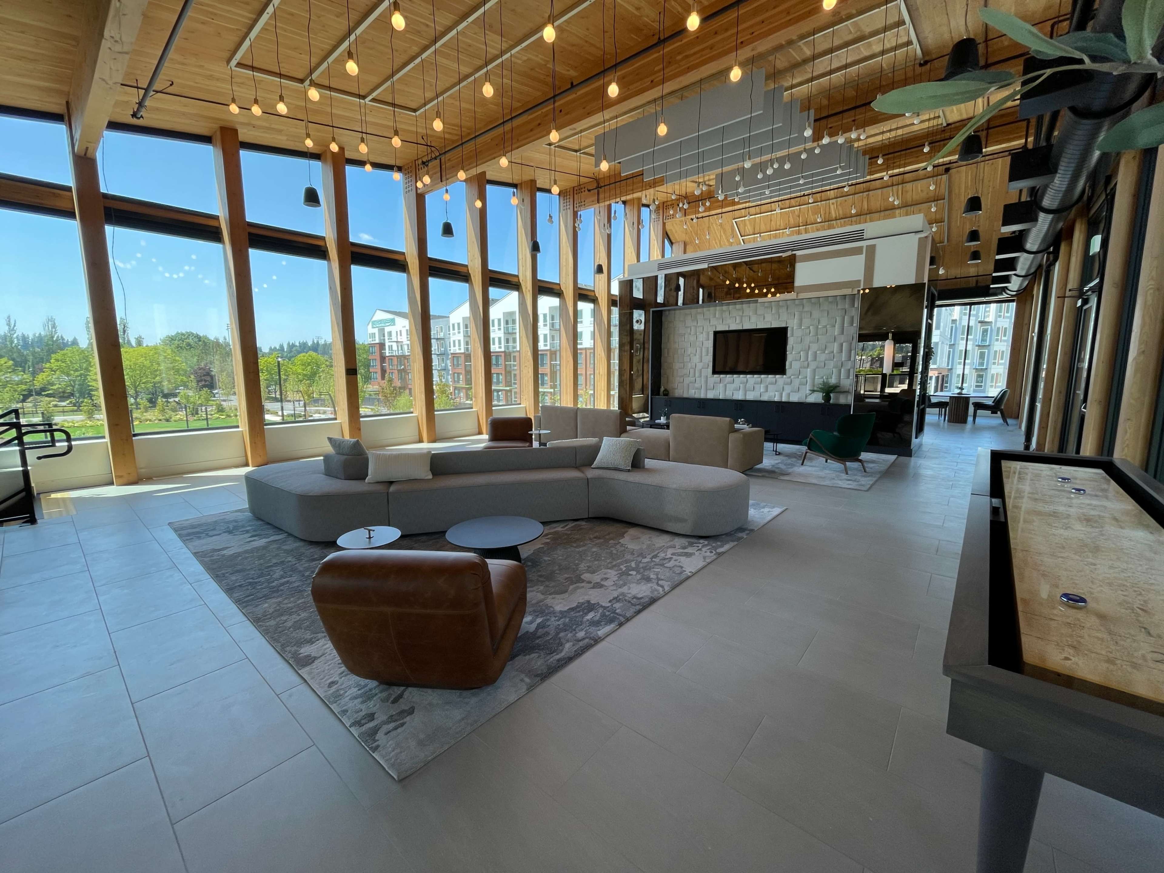 A spacious, modern lounge area with large windows, wooden beams, and a combination of sofas and chairs arranged around a central coffee table.