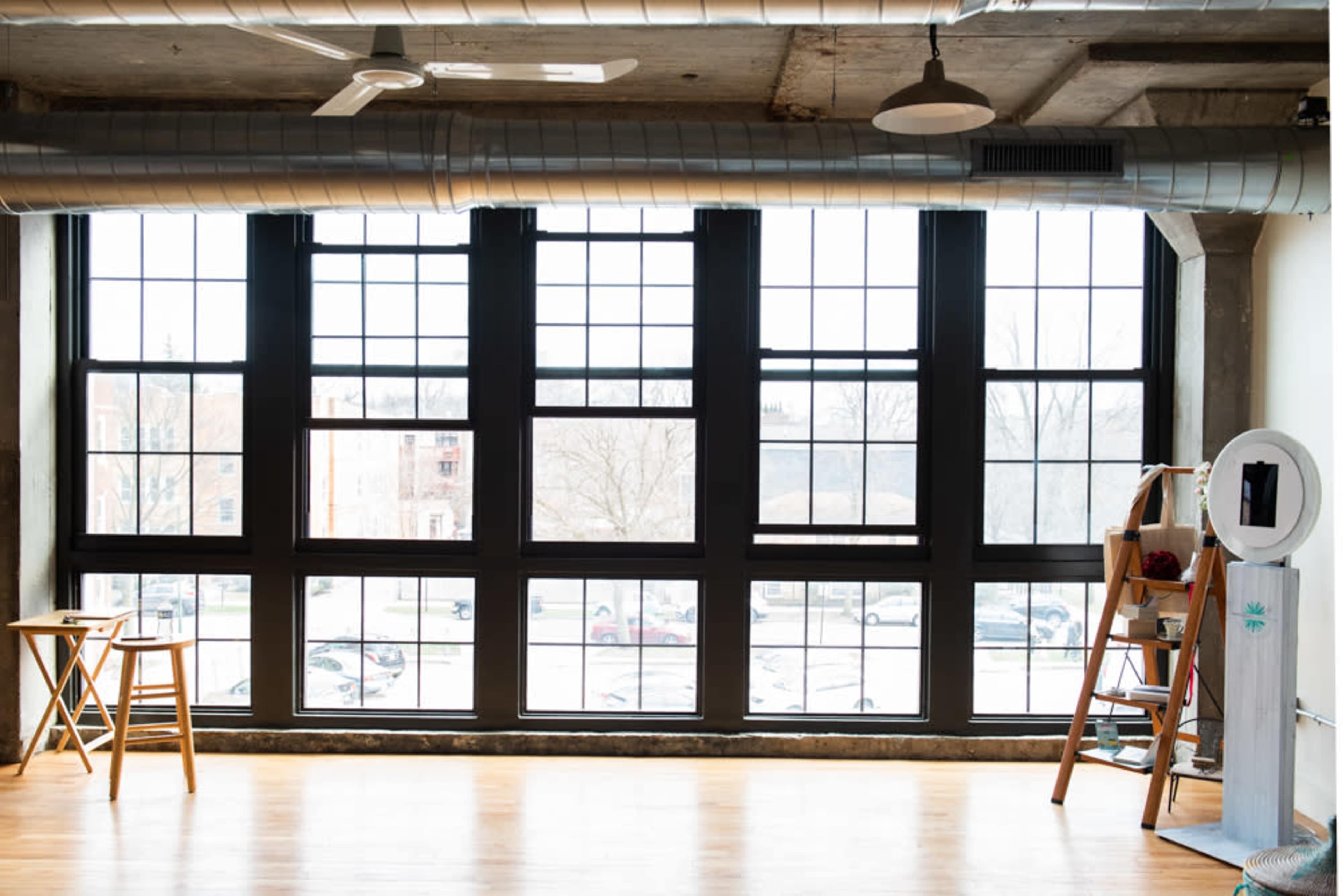 A spacious room with large black-framed windows overlooking a parking area, featuring a folding table, a ladder, and a round mirror.