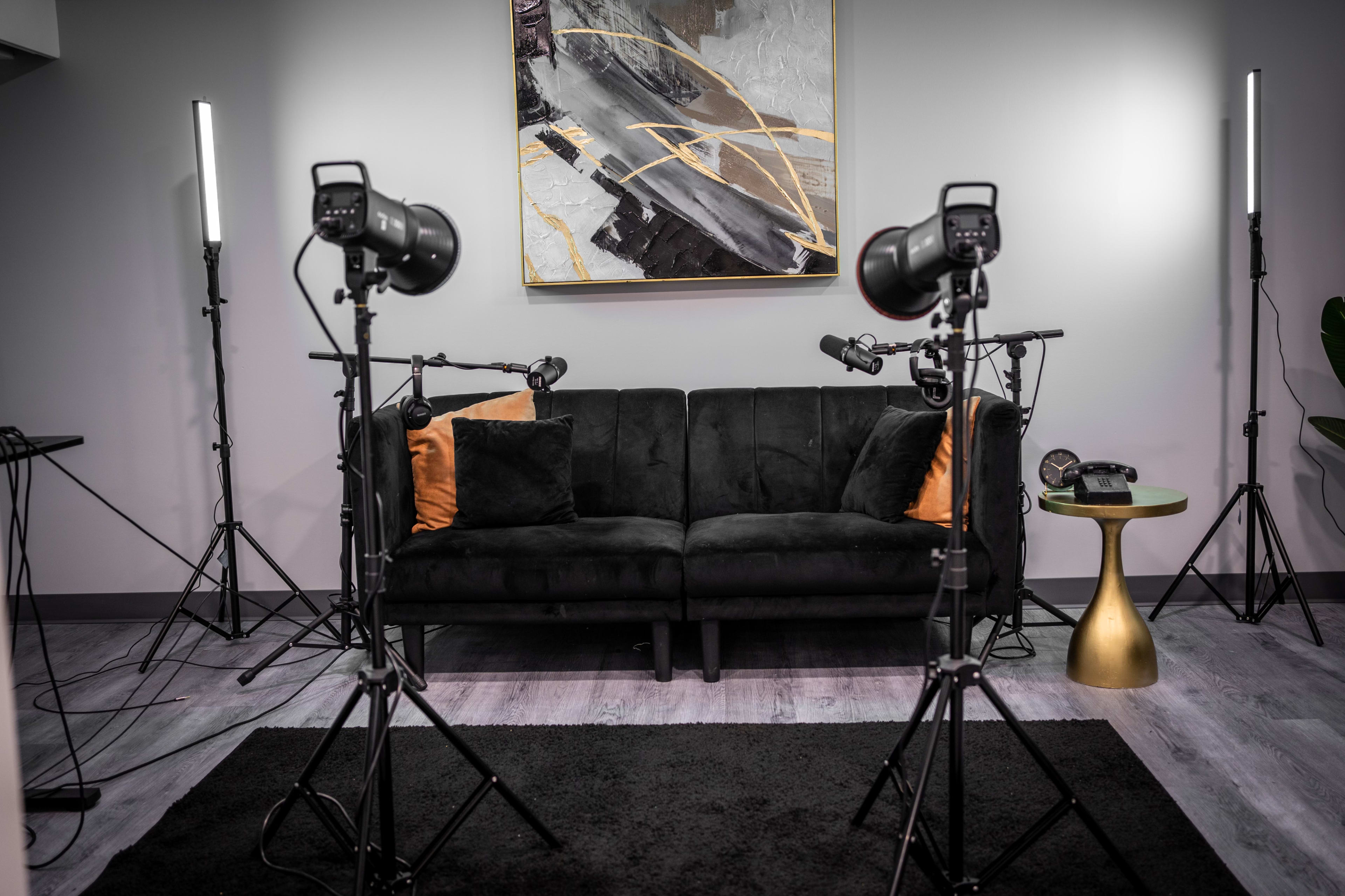 A modern interview setup featuring a black sofa with two pillows, flanked by tripods holding lights and microphones, against a grey wall adorned with abstract artwork.