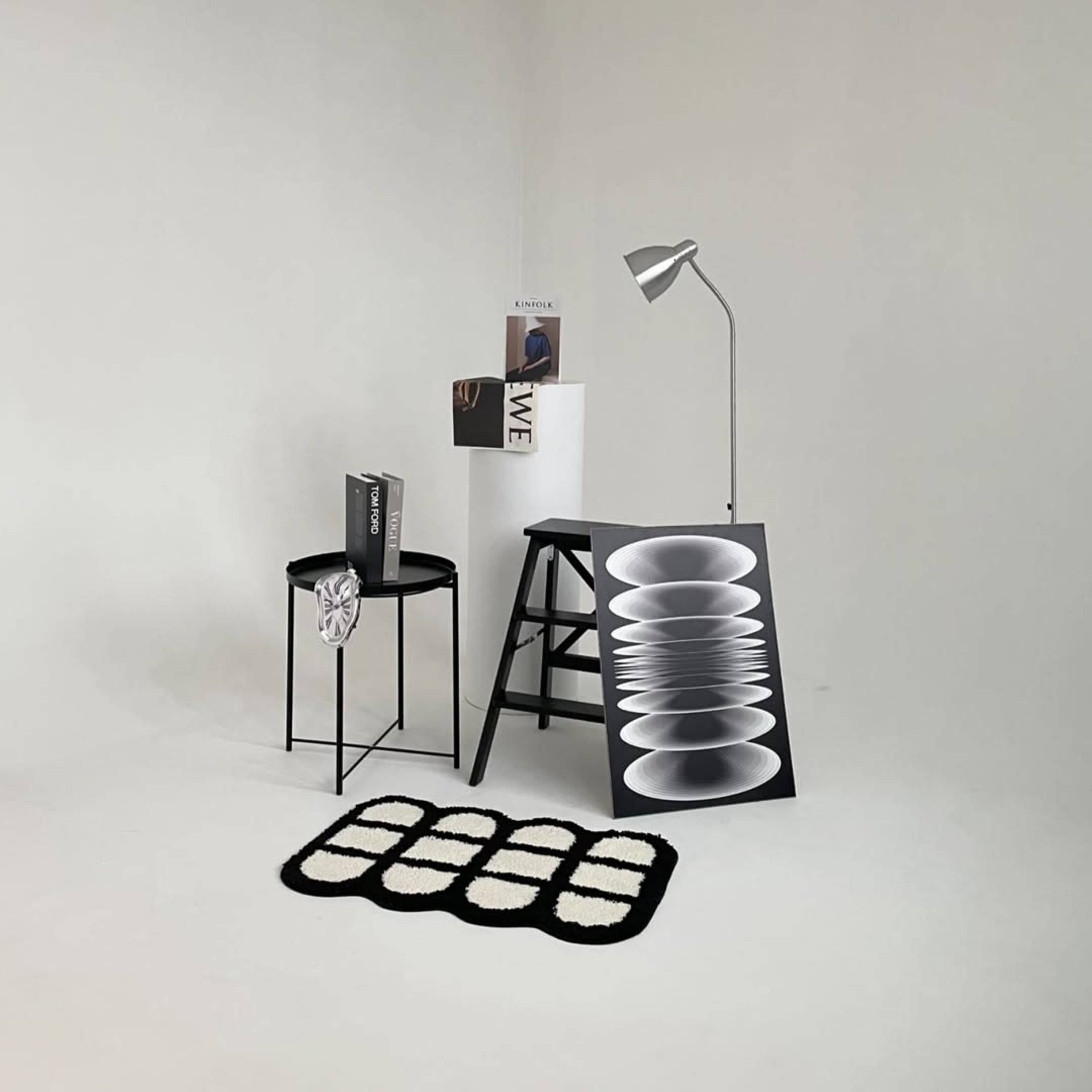 The scene features a minimalist setup with a black stool, a white lamp, a ladder-like table displaying an artwork, and stacked books on a bare white floor.