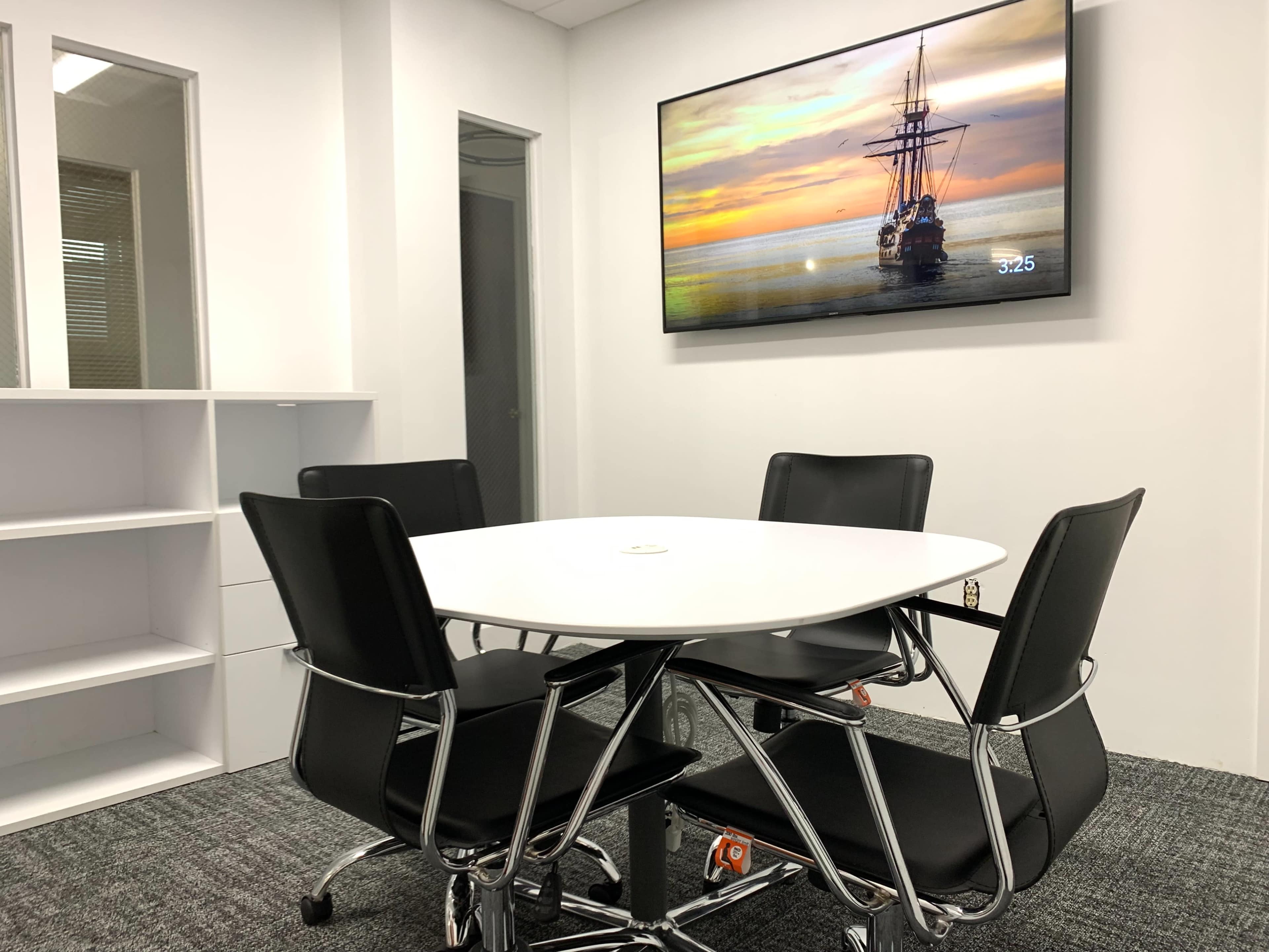 A modern conference room features a round table surrounded by black chairs and a television displaying a ship on the wall.