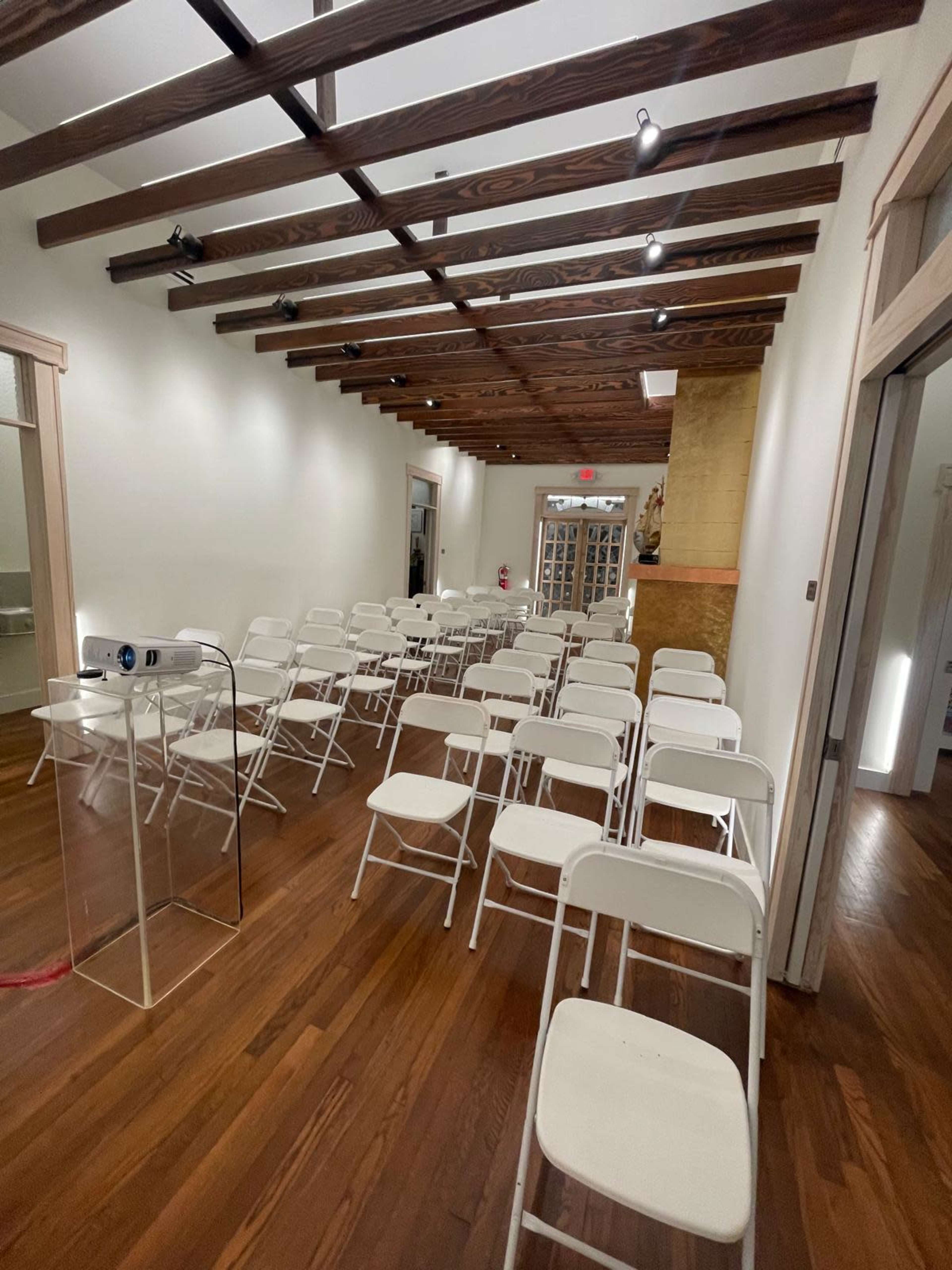 A row of white folding chairs arranged in a well-lit room with wooden beams on the ceiling and polished wooden floors.