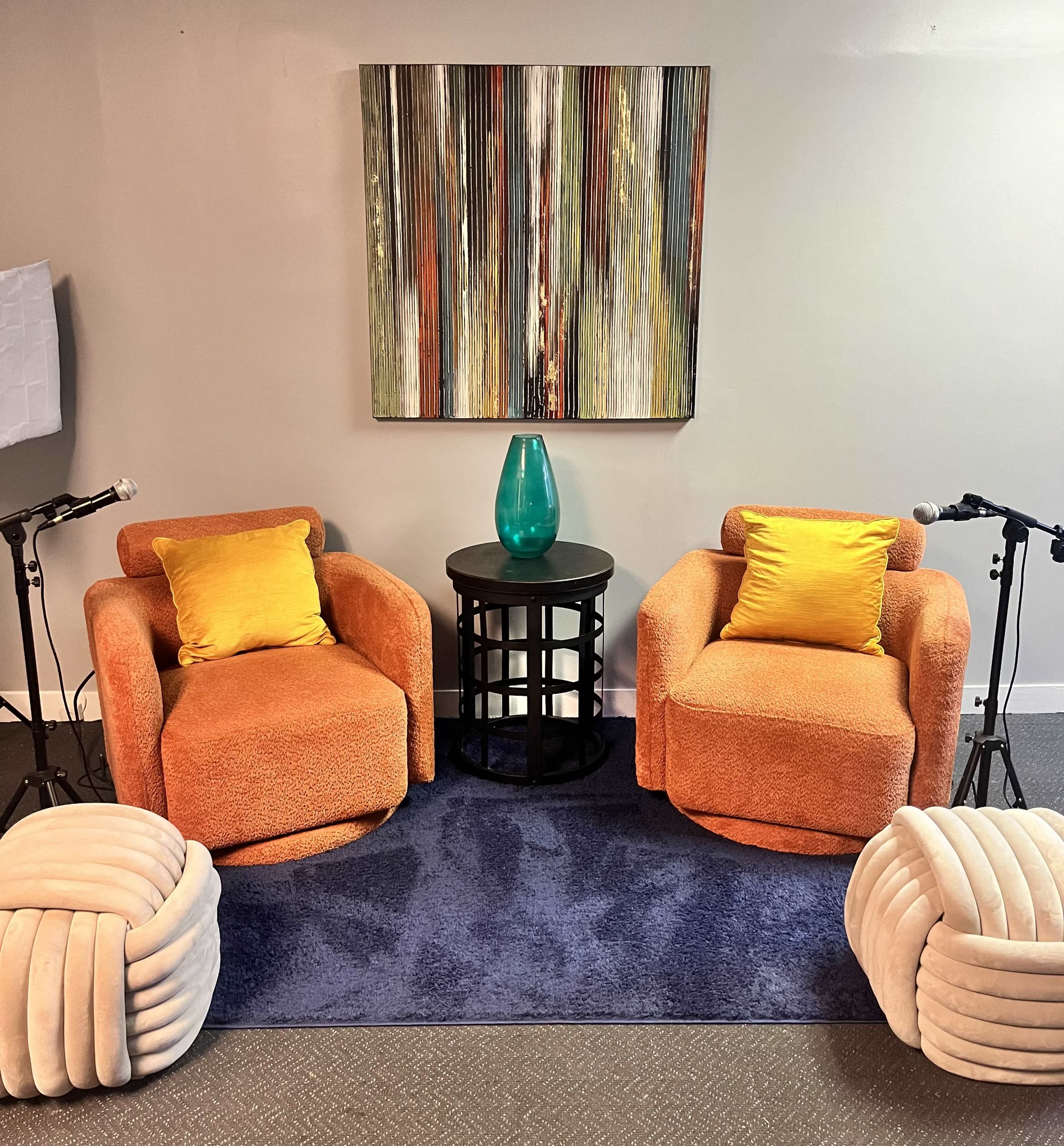 A cozy seating area with two orange armchairs, a blue rug underneath, a round black table with a teal vase, and two microphones positioned nearby.