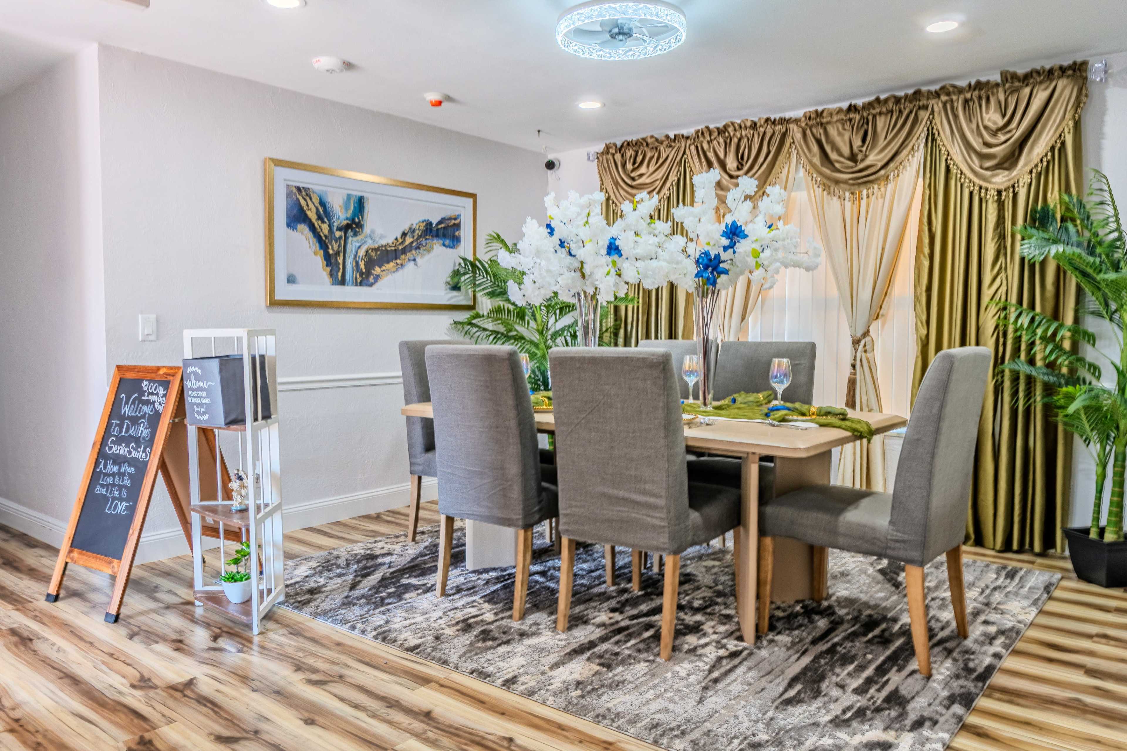 A dining room features a wooden table set for six, surrounded by gray upholstered chairs, with decorative plants and a large floral centerpiece.
