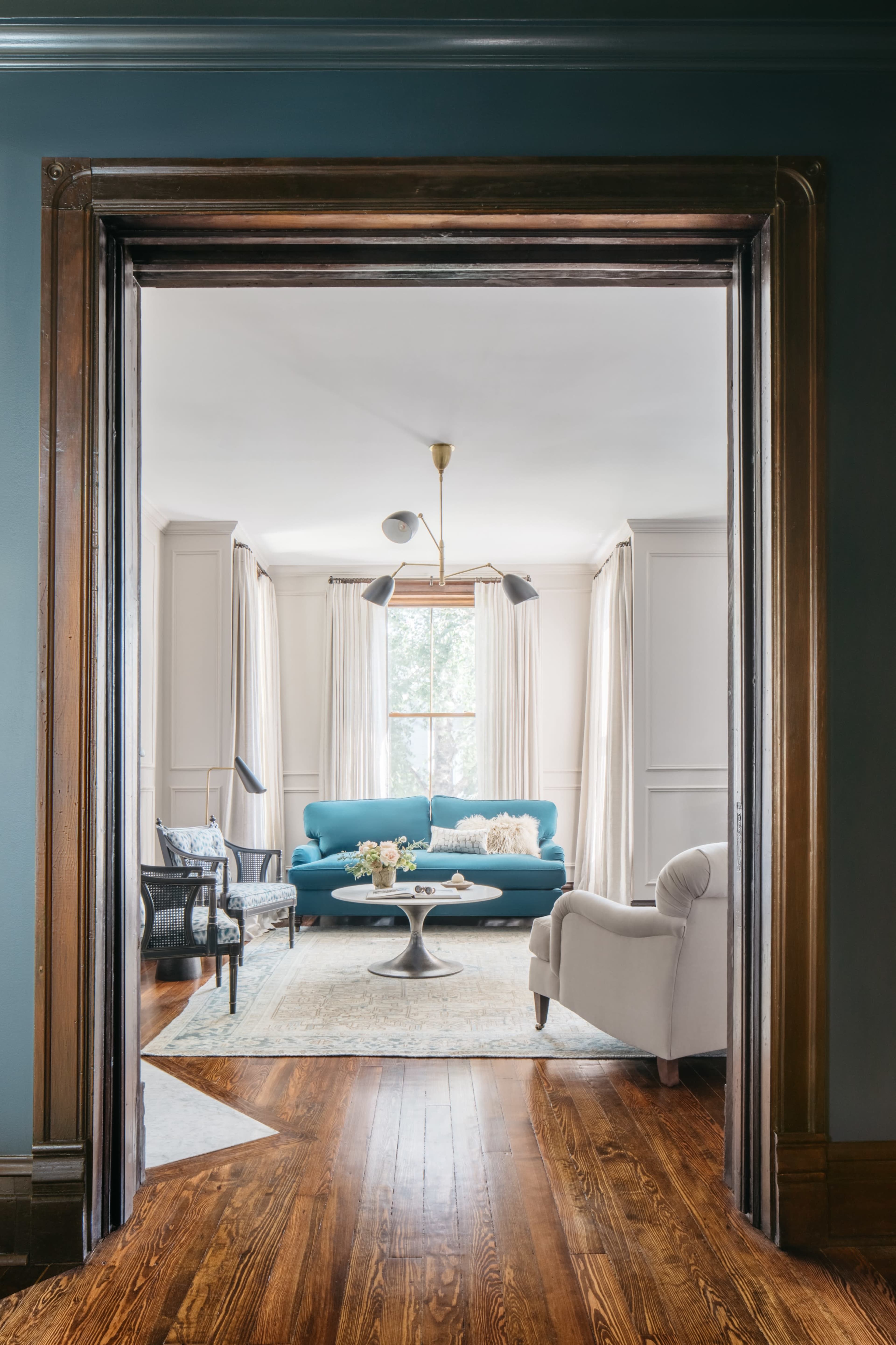 A view through a wooden archway reveals a bright living room with a blue sofa, white armchair, and large windows.