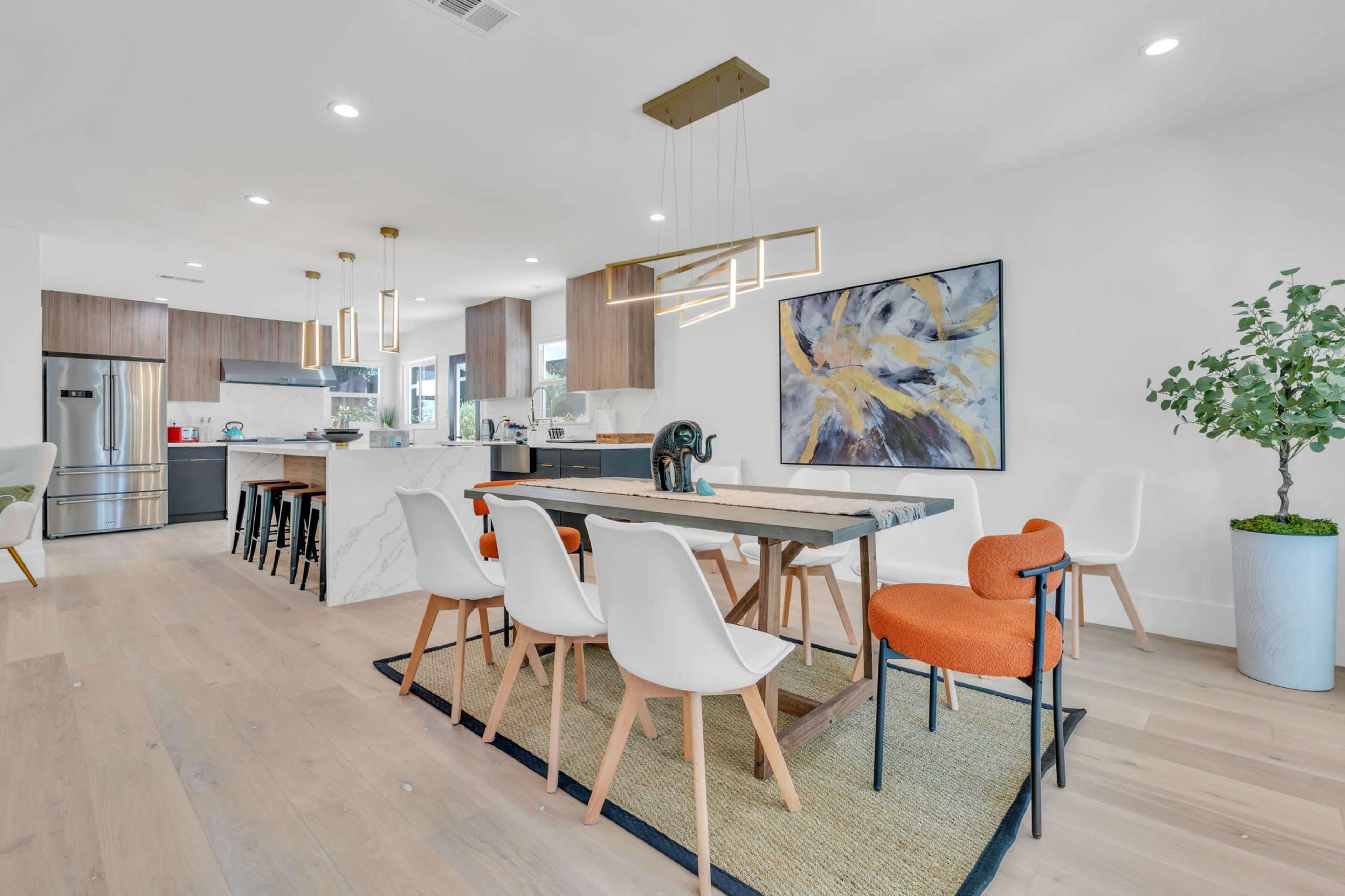 A modern dining area features a rectangular table with white and orange chairs, surrounded by light wood flooring and open kitchen cabinetry.