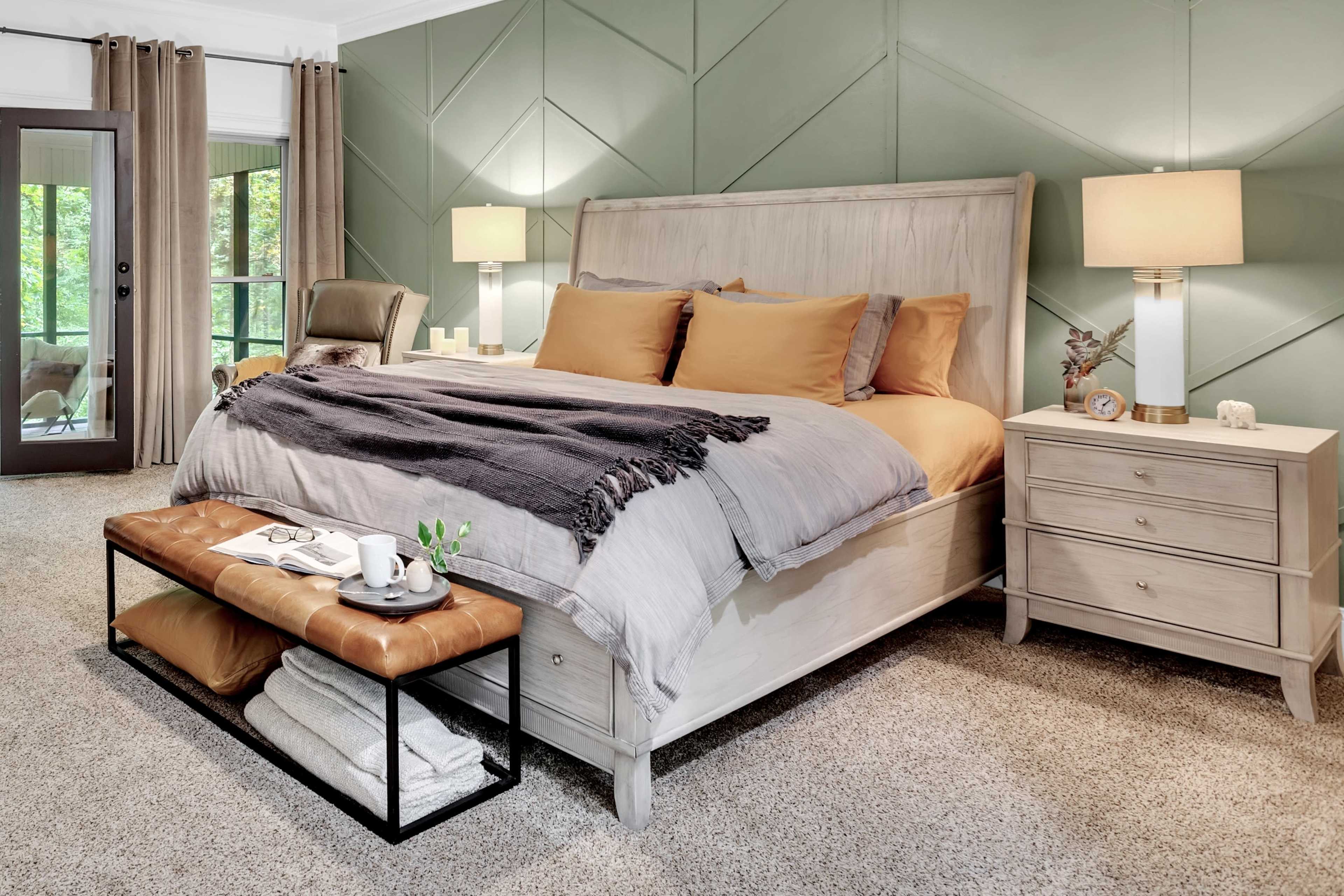 The image depicts a well-furnished bedroom featuring a large bed with orange and gray bedding, a wooden nightstand, and a bench at the foot of the bed, all set against a green accent wall.