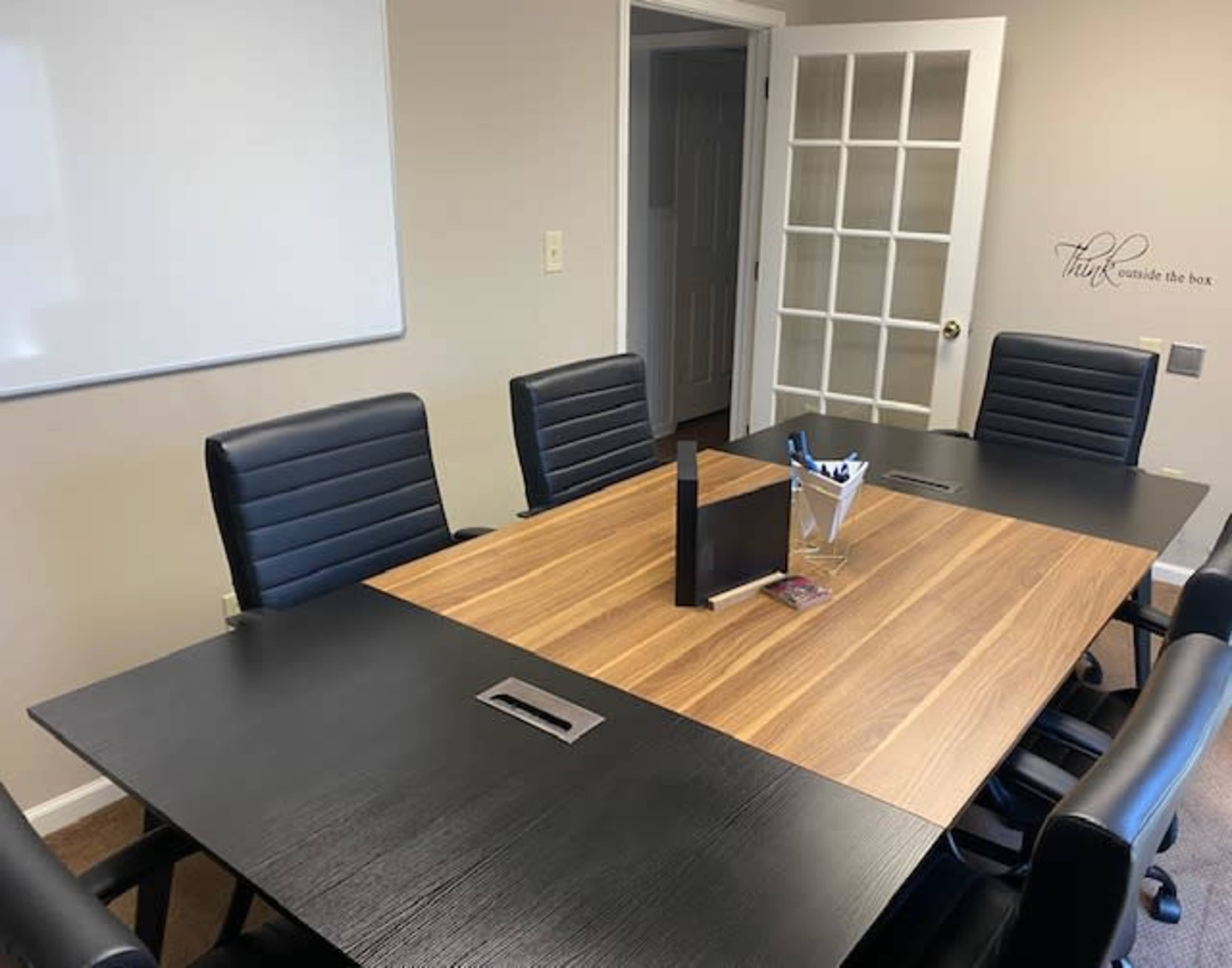 A modern conference room features a wooden table surrounded by black leather chairs, with a whiteboard and a glass door in the background.