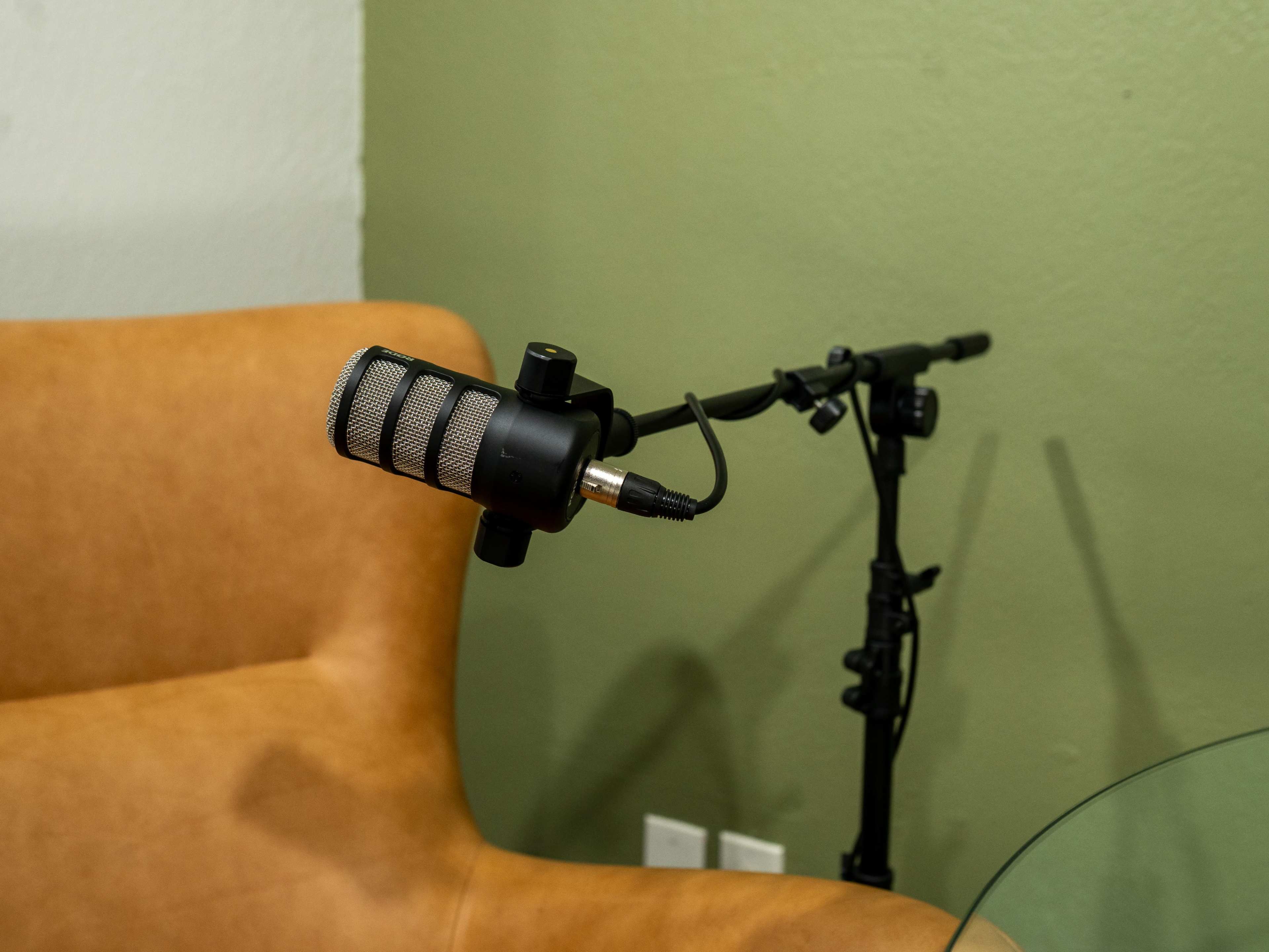 A microphone is mounted on a stand next to a brown sofa and a glass table against a green wall.