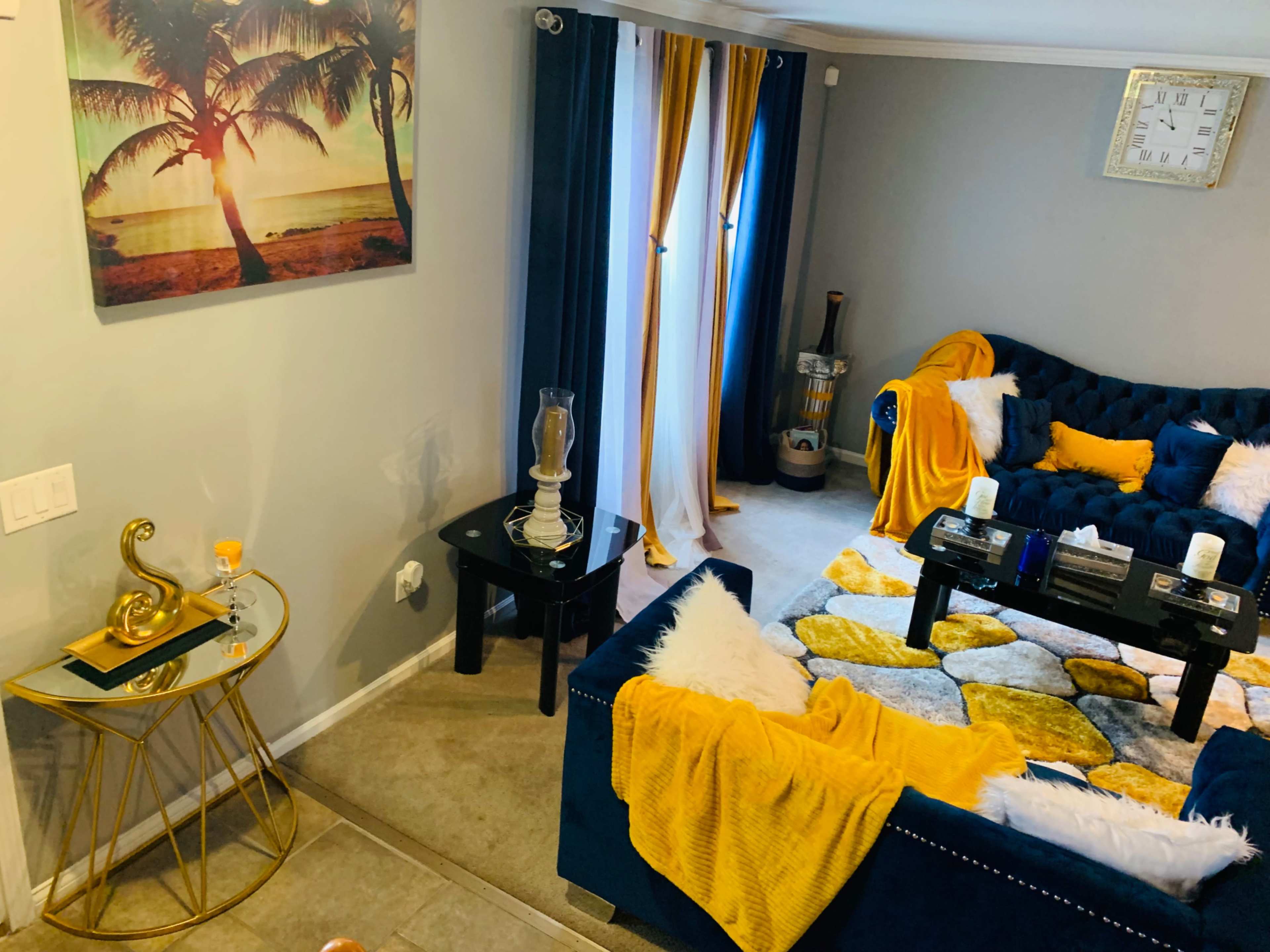 A living room with a blue sofa, a coffee table, a patterned rug, and decorative elements including curtains, a wall clock, and palm tree artwork.