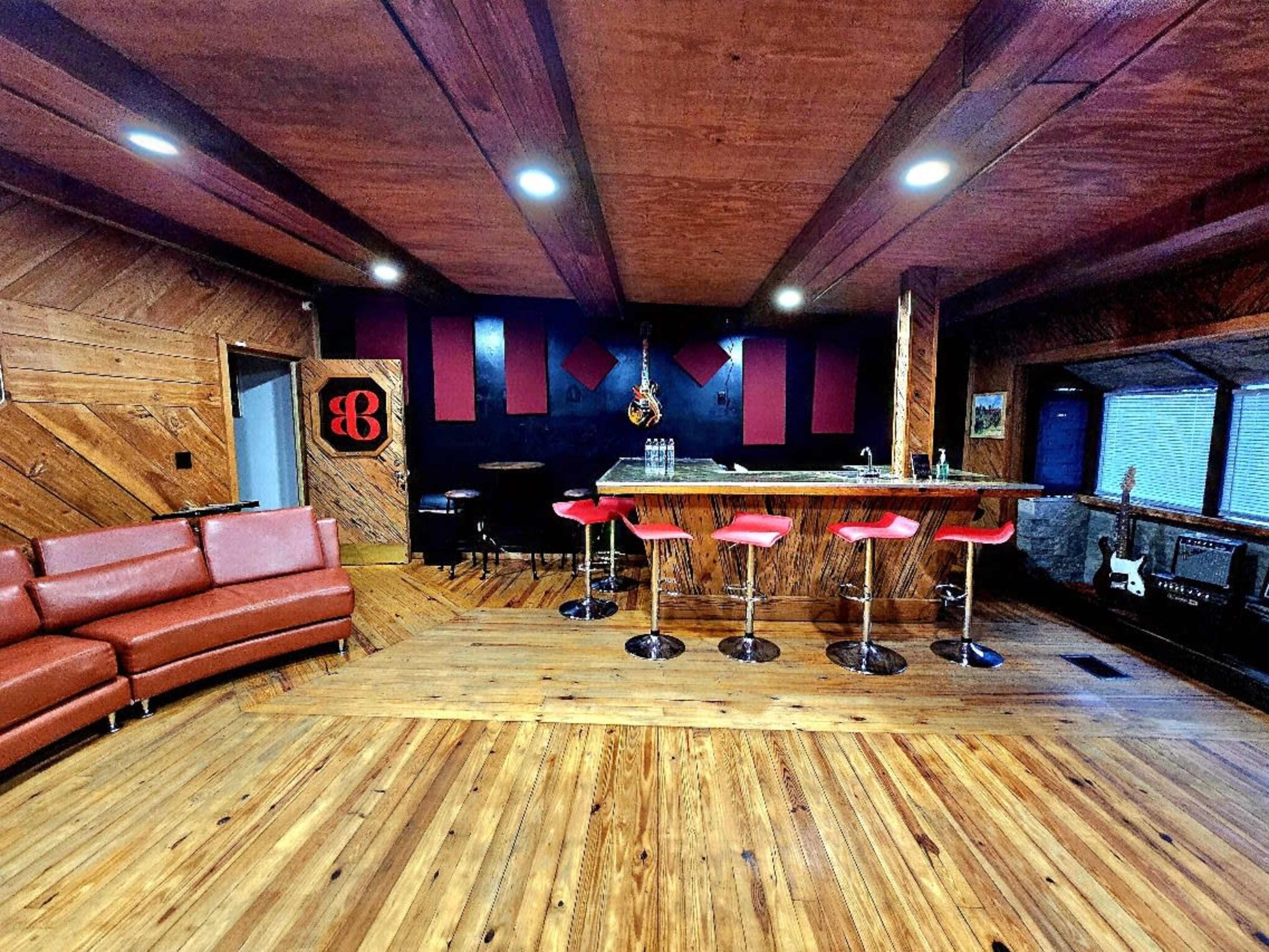 The image shows a spacious room featuring a bar with red stools, a large leather sectional couch, and wooden flooring, accented by dark walls and decorative elements.