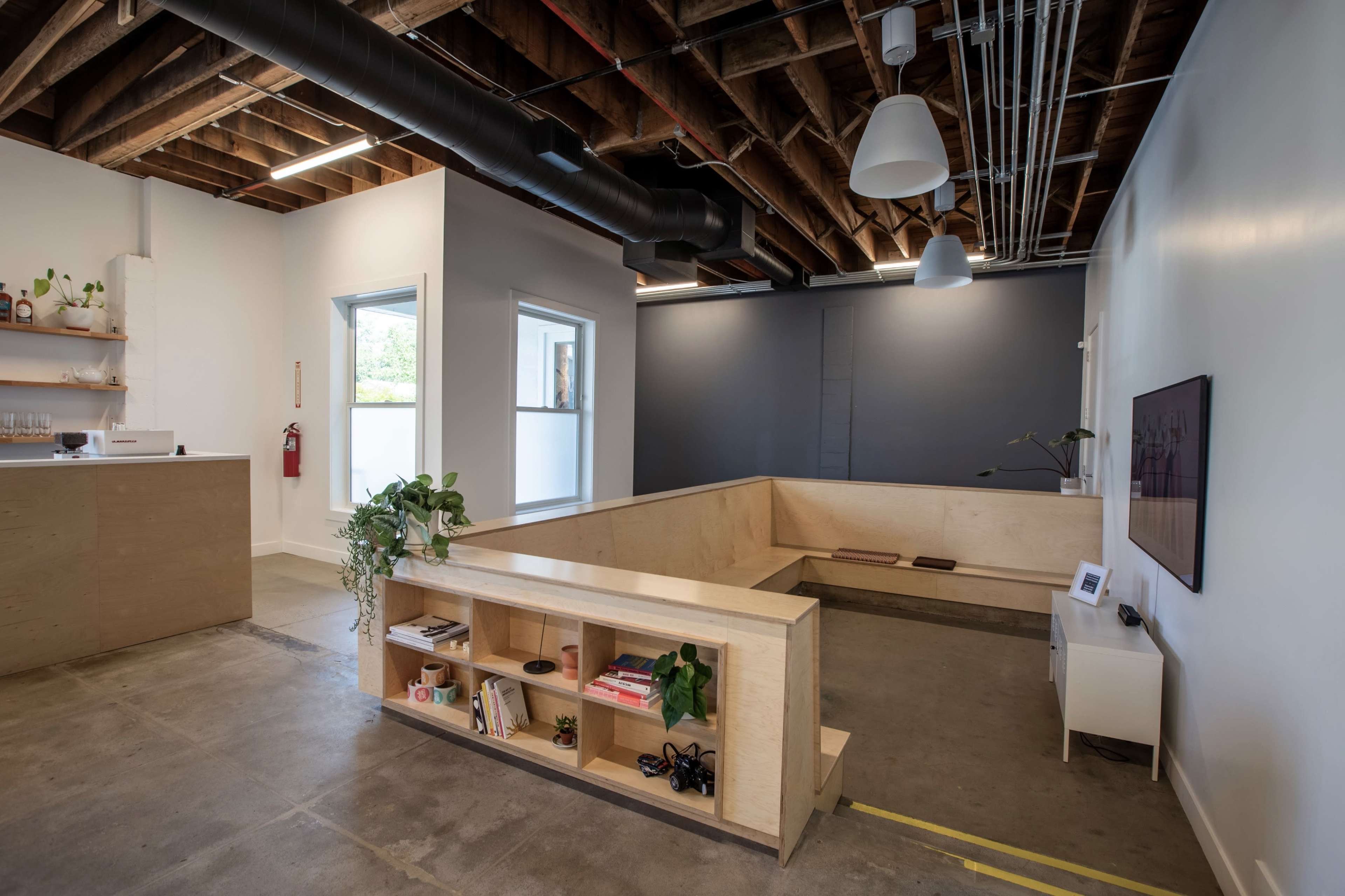A modern office space with exposed wooden beams, minimalist furniture, and a low wooden seating area surrounded by large windows.