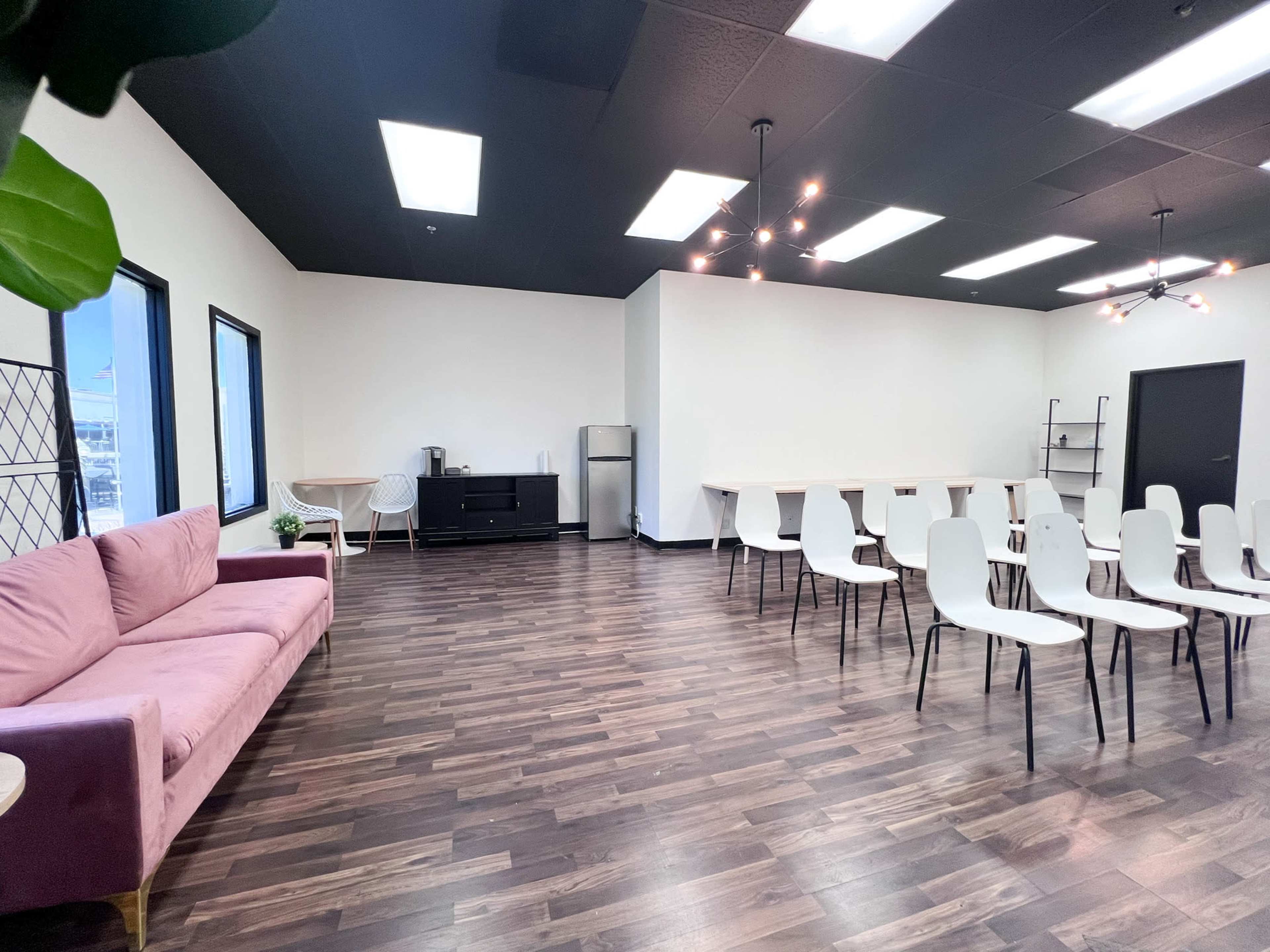 A spacious, modern meeting room with a pink sofa, a black cabinet, a refrigerator, and rows of white chairs arranged around a central table space.