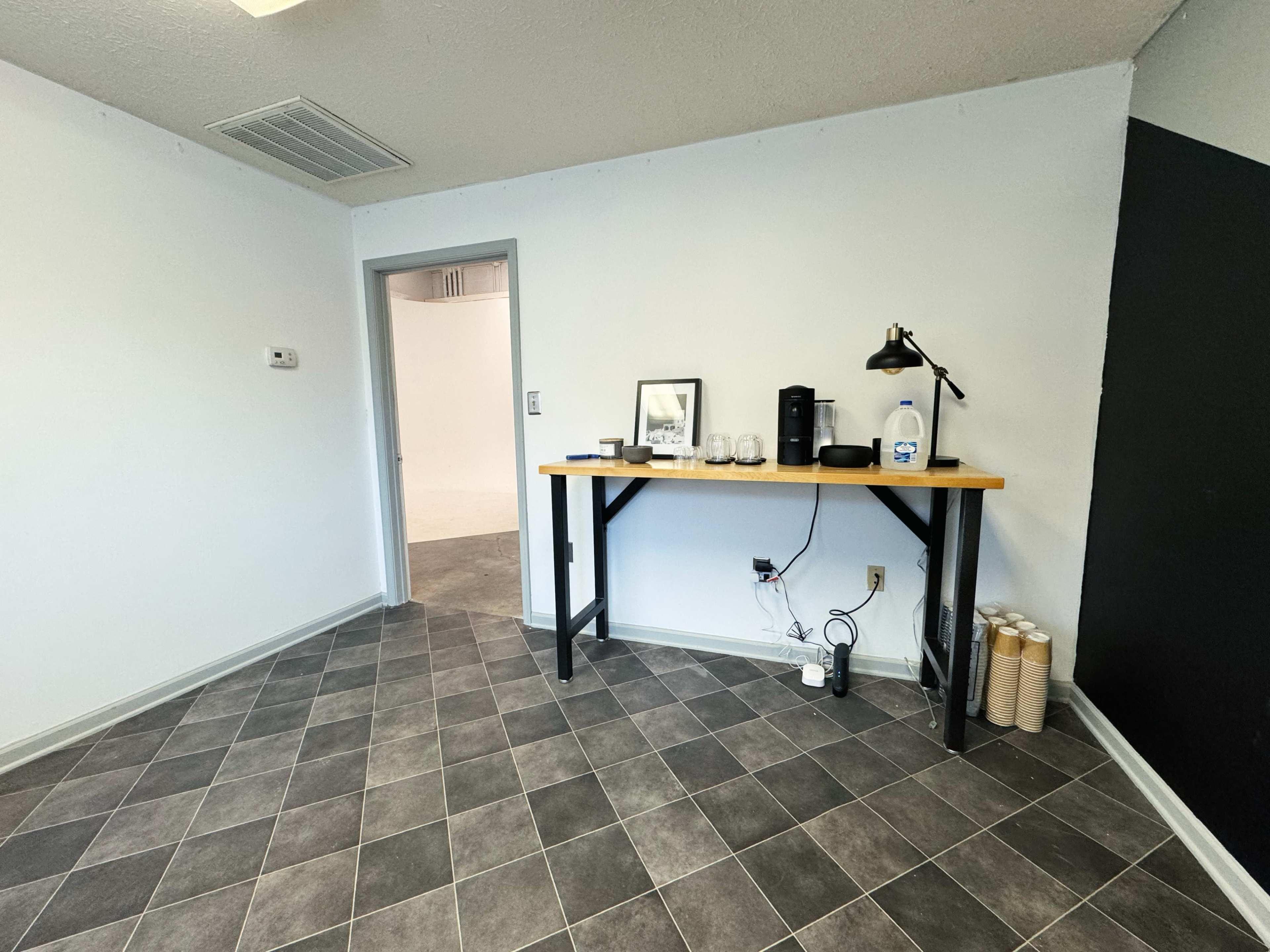 A light-colored room with a gray tiled floor, featuring a table with a coffee maker, utensils, and a lamp near an entrance door.