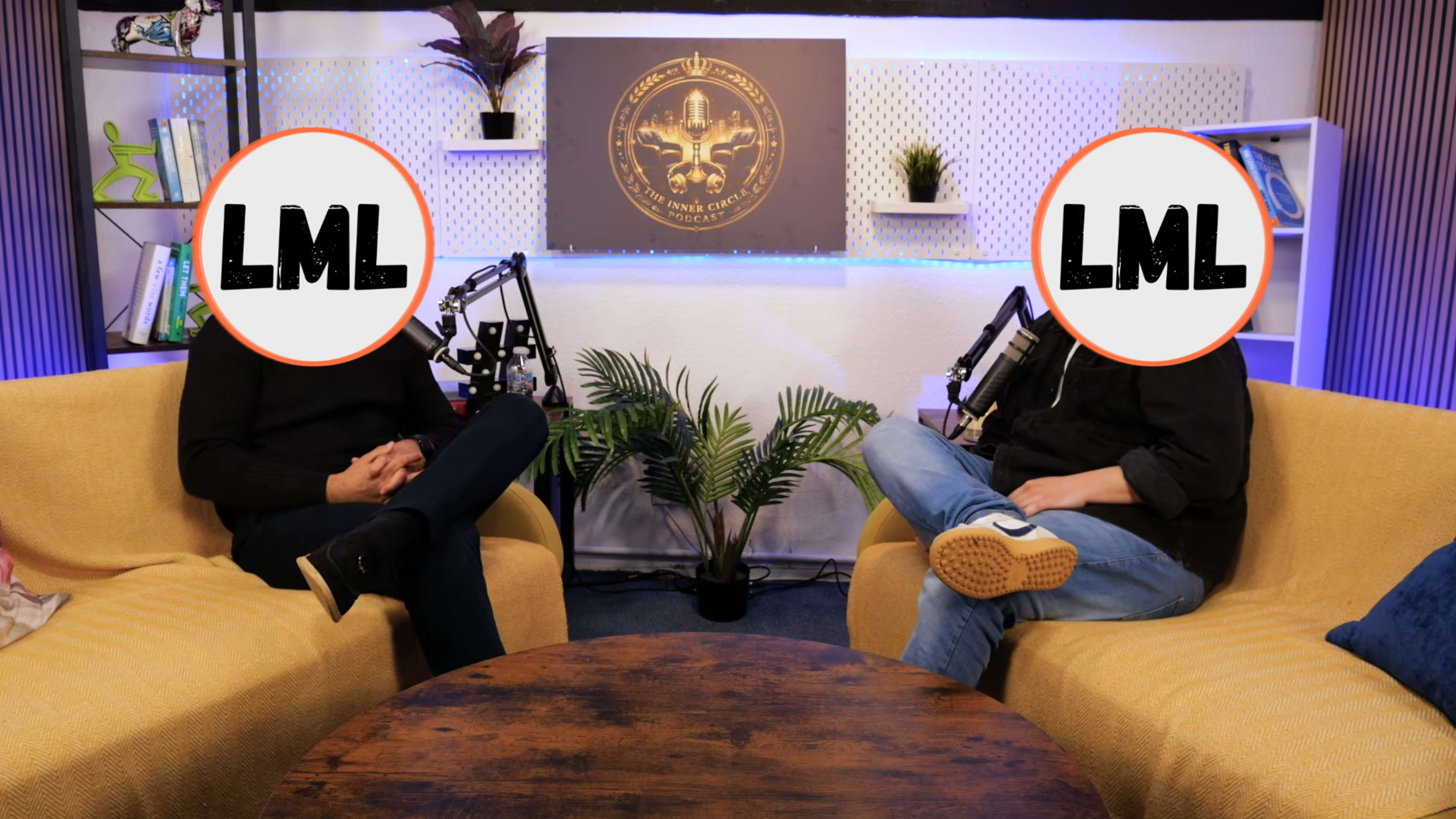 Two individuals sit in yellow chairs across from each other at a round wooden table in a studio setting, with microphones positioned nearby and a plant in the background.
