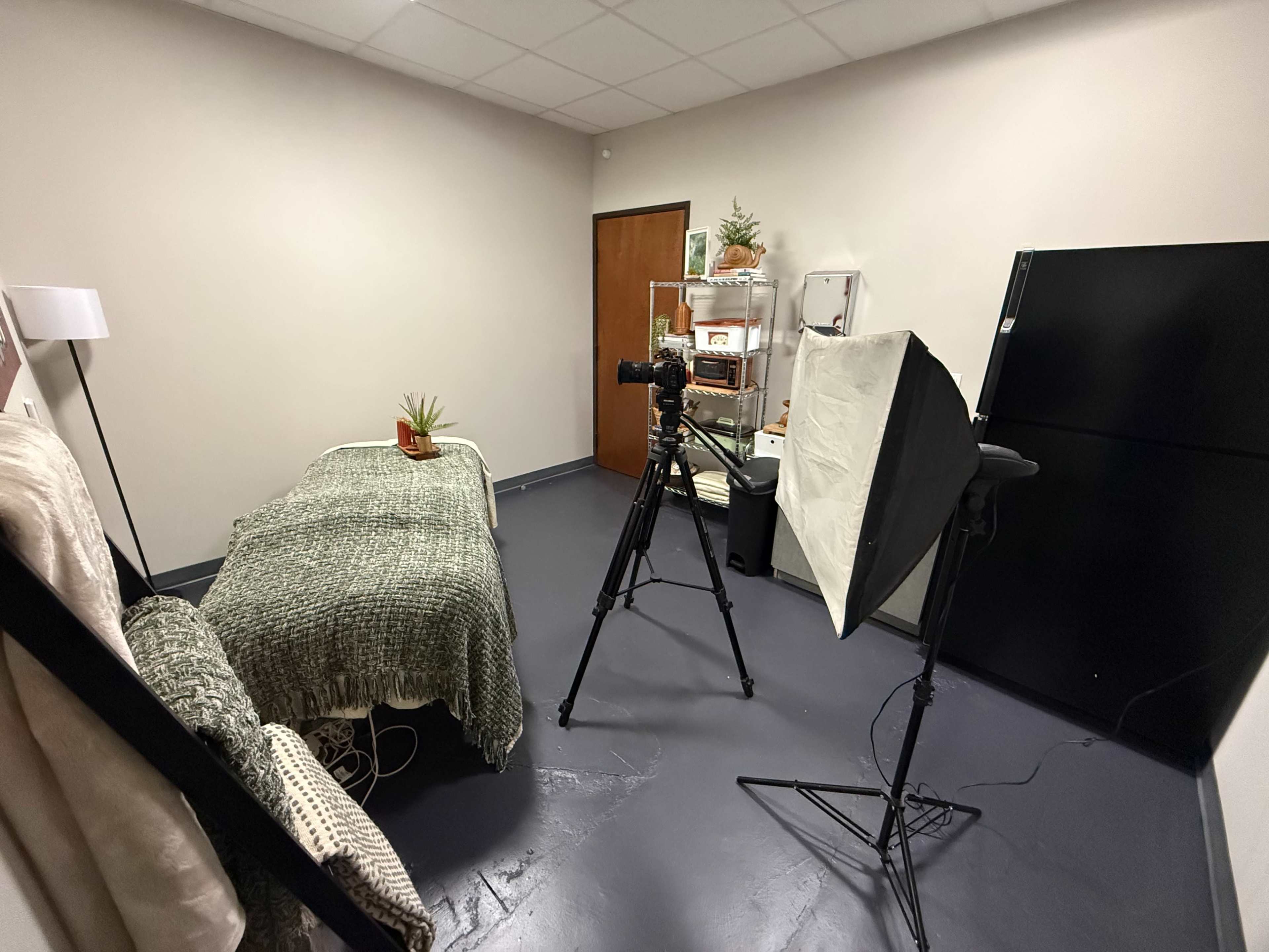 The image shows a small room with a bed covered by a green blanket, a camera on a tripod, and a light setup next to a black refrigerator.