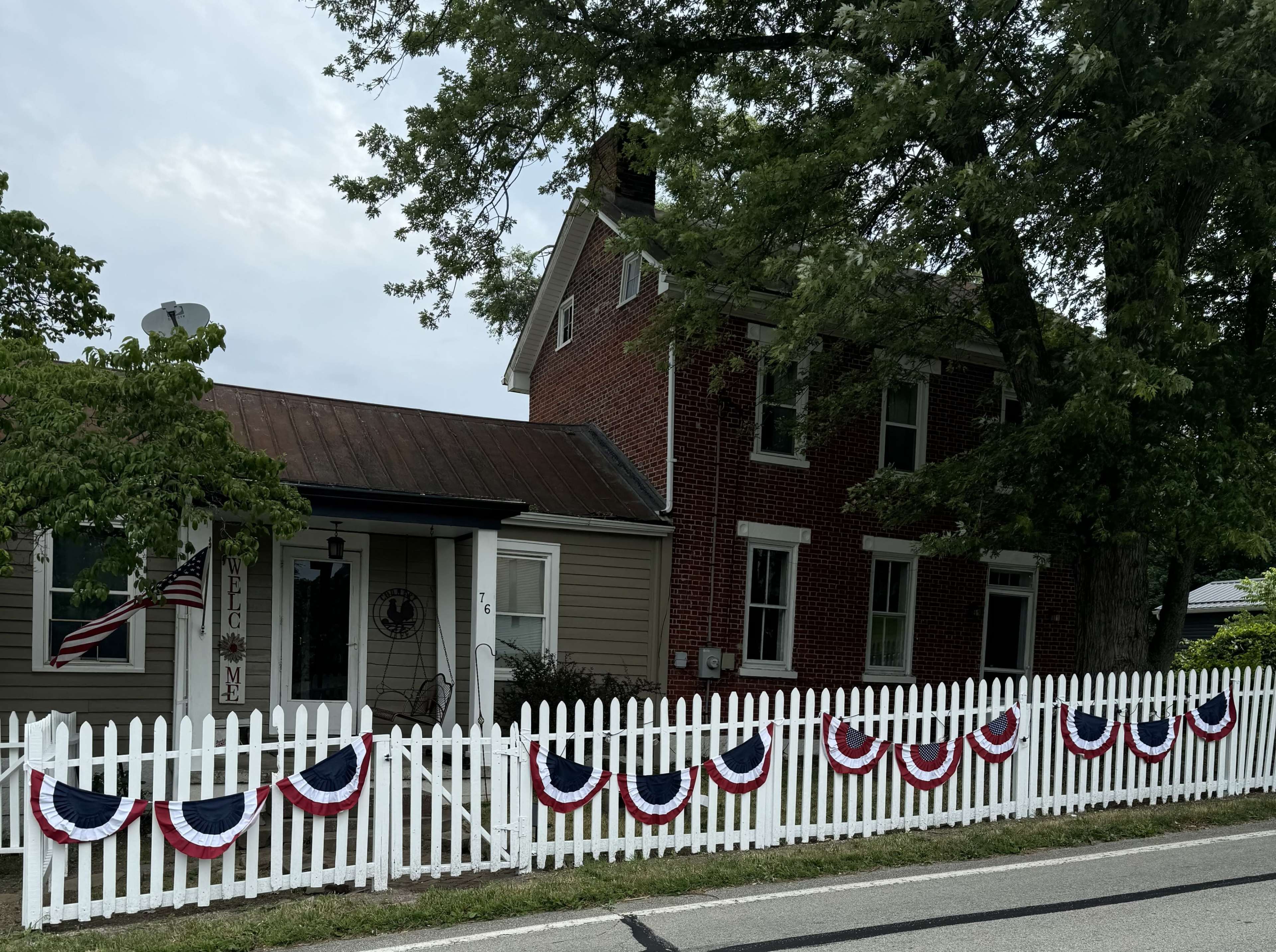 A two-story brick house with a covered porch is adorned with red, white, and blue bunting, and a white picket fence runs along the front.