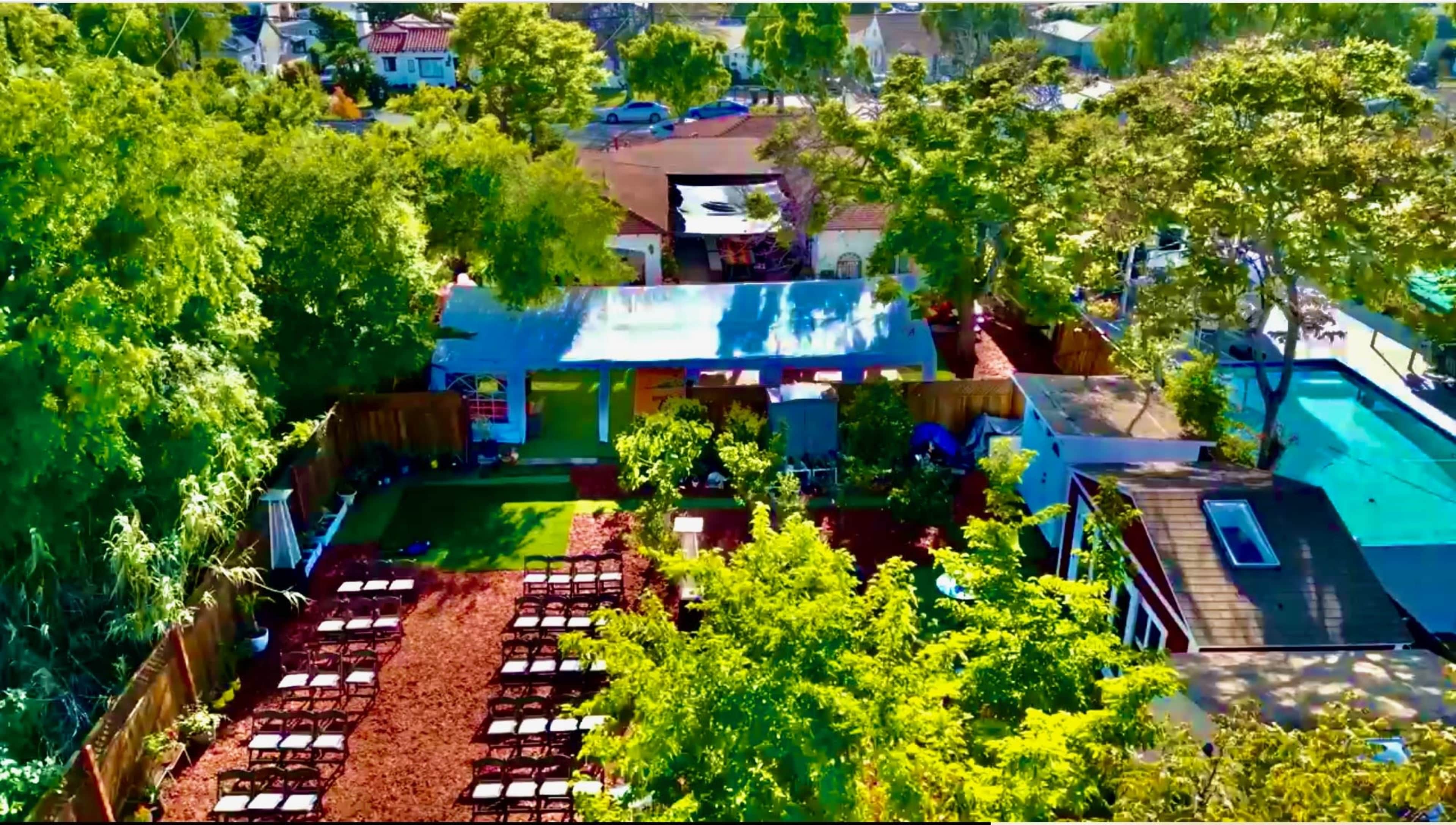 The image shows a lush garden area with arranged seating, surrounded by trees and small buildings in a residential neighborhood.