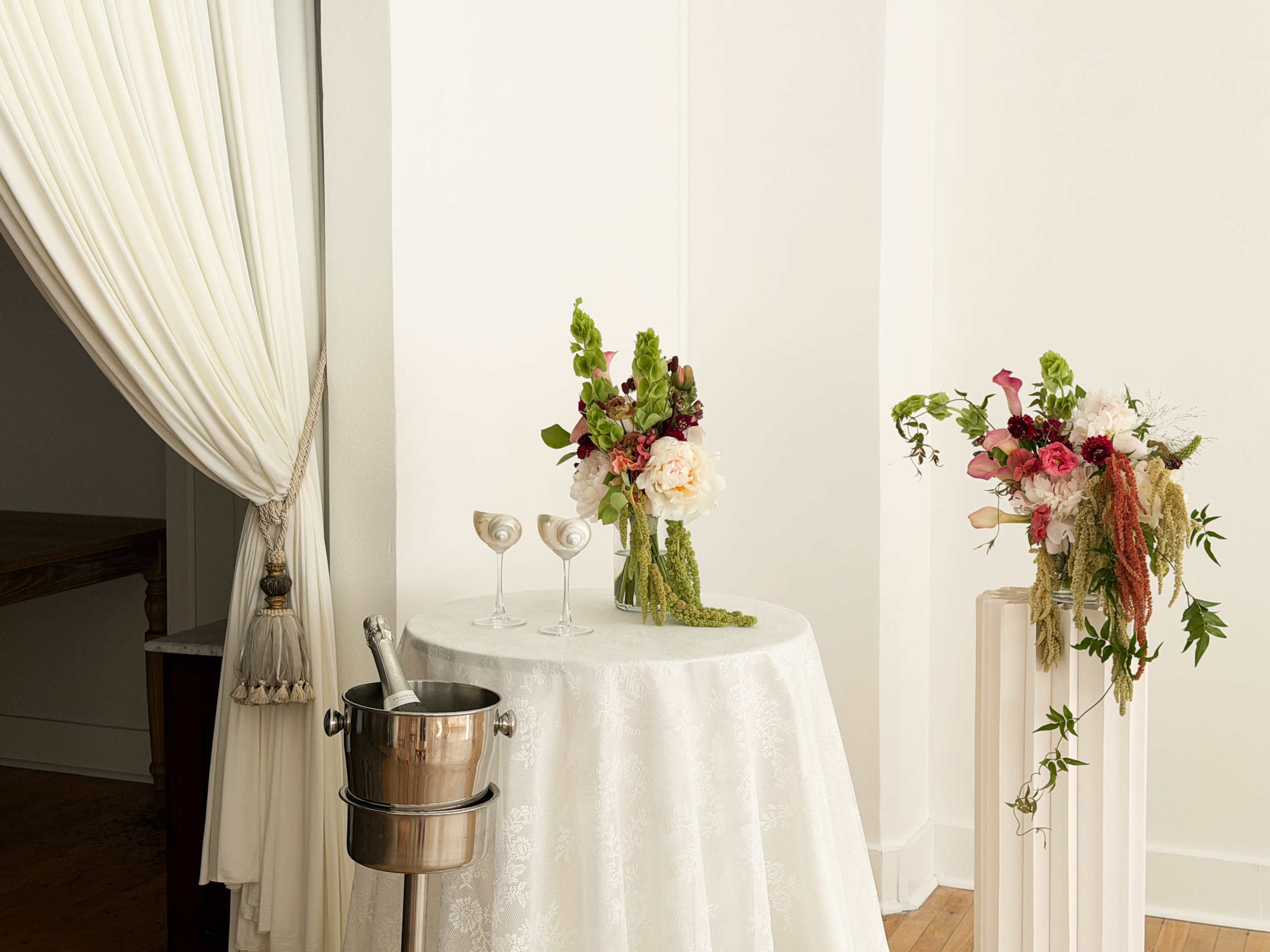 A round table with a white cloth displays two champagne glasses, a chilled champagne bucket, and two floral arrangements in vases on either side.