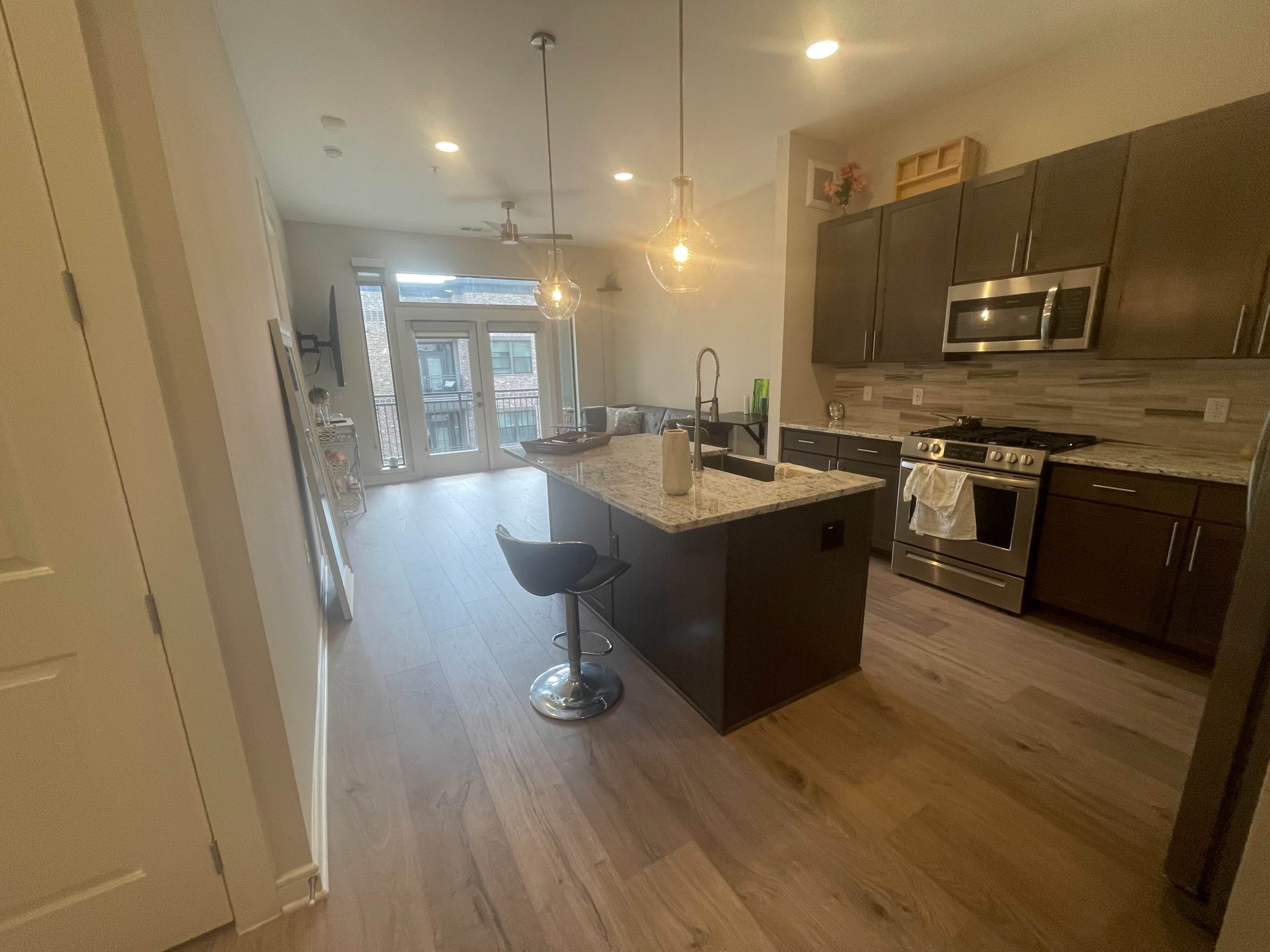 The image shows a modern kitchen with granite countertops, dark cabinetry, a stainless steel stove, and a small dining area featuring a bar stool.