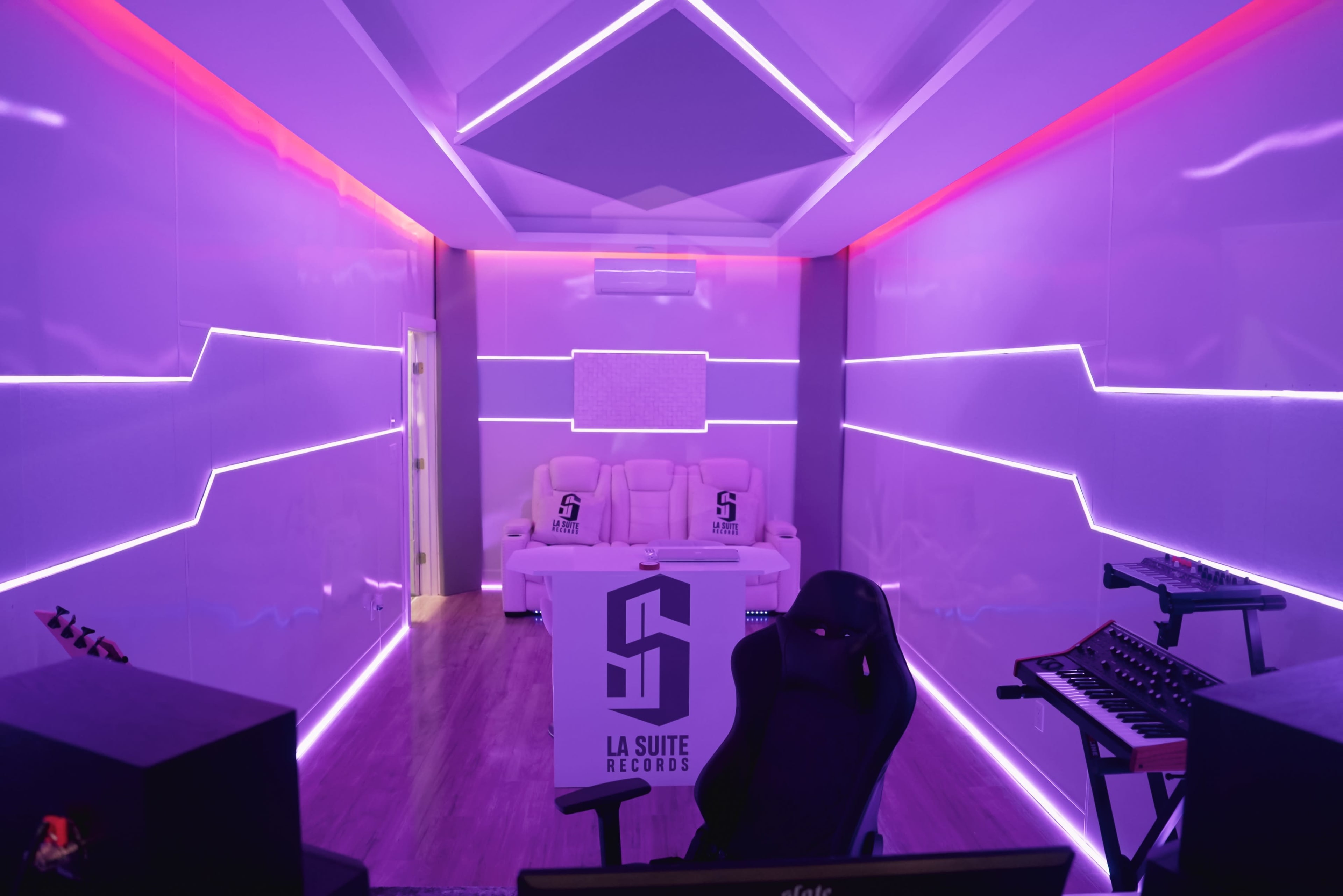 A modern recording studio with purple LED light accents, featuring a seating area, a desk with music equipment, and sleek walls.