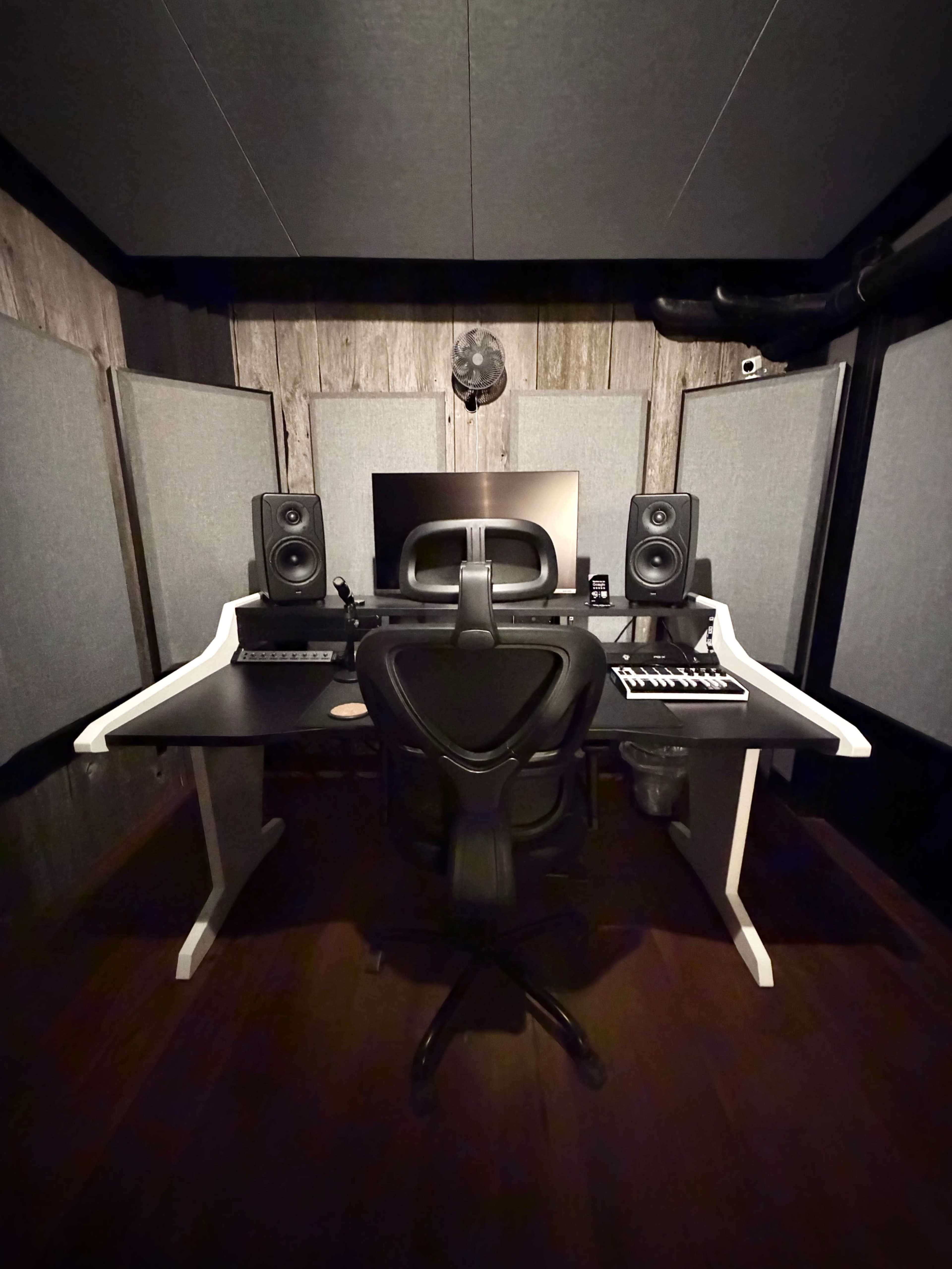 The image shows a music production studio featuring a desk with studio monitors, a computer, a MIDI keyboard, and soundproofing panels on the walls.