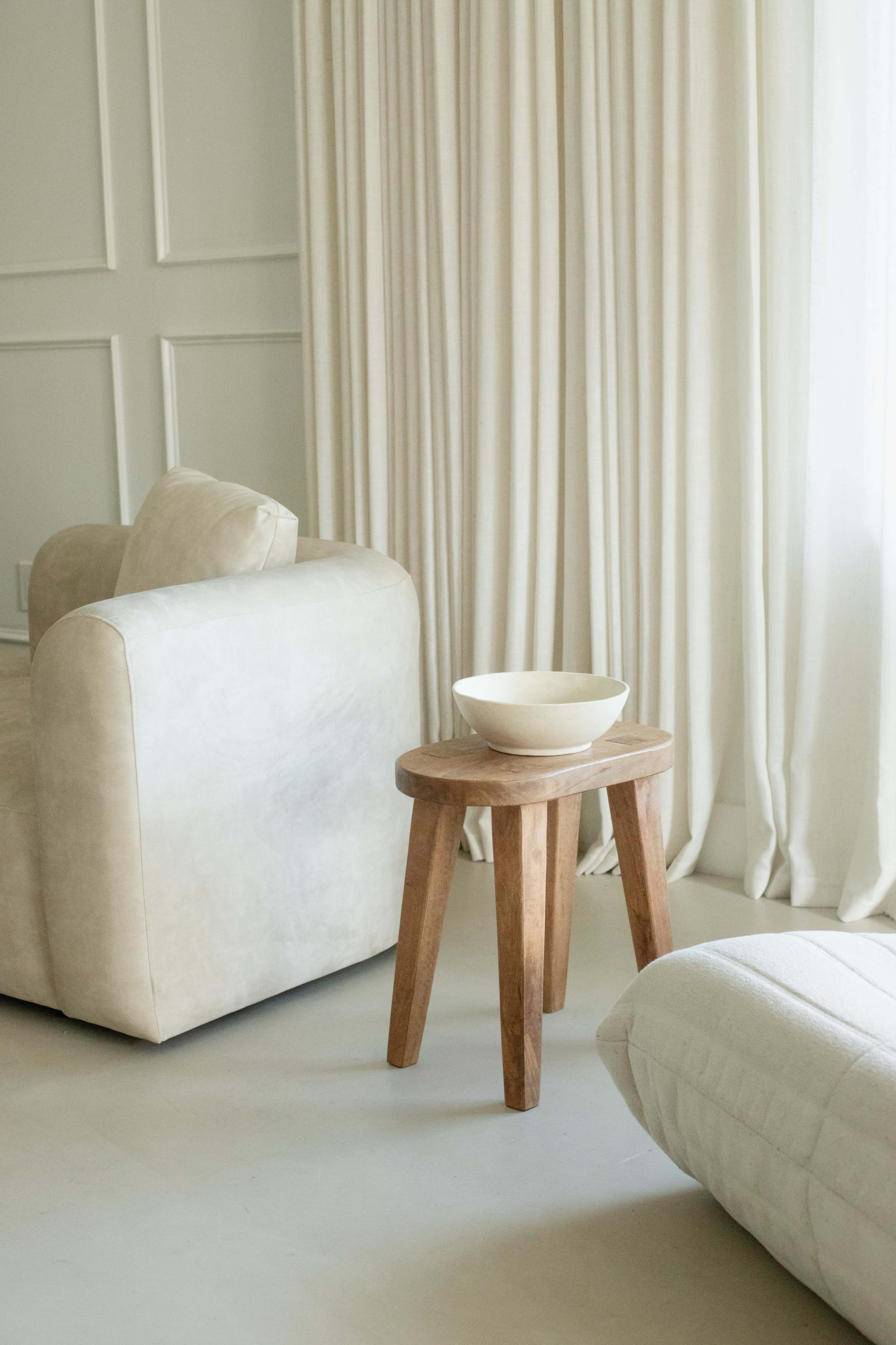 The image features a light-colored living room with a beige couch and a wooden side table holding a bowl, beside sheer white curtains.