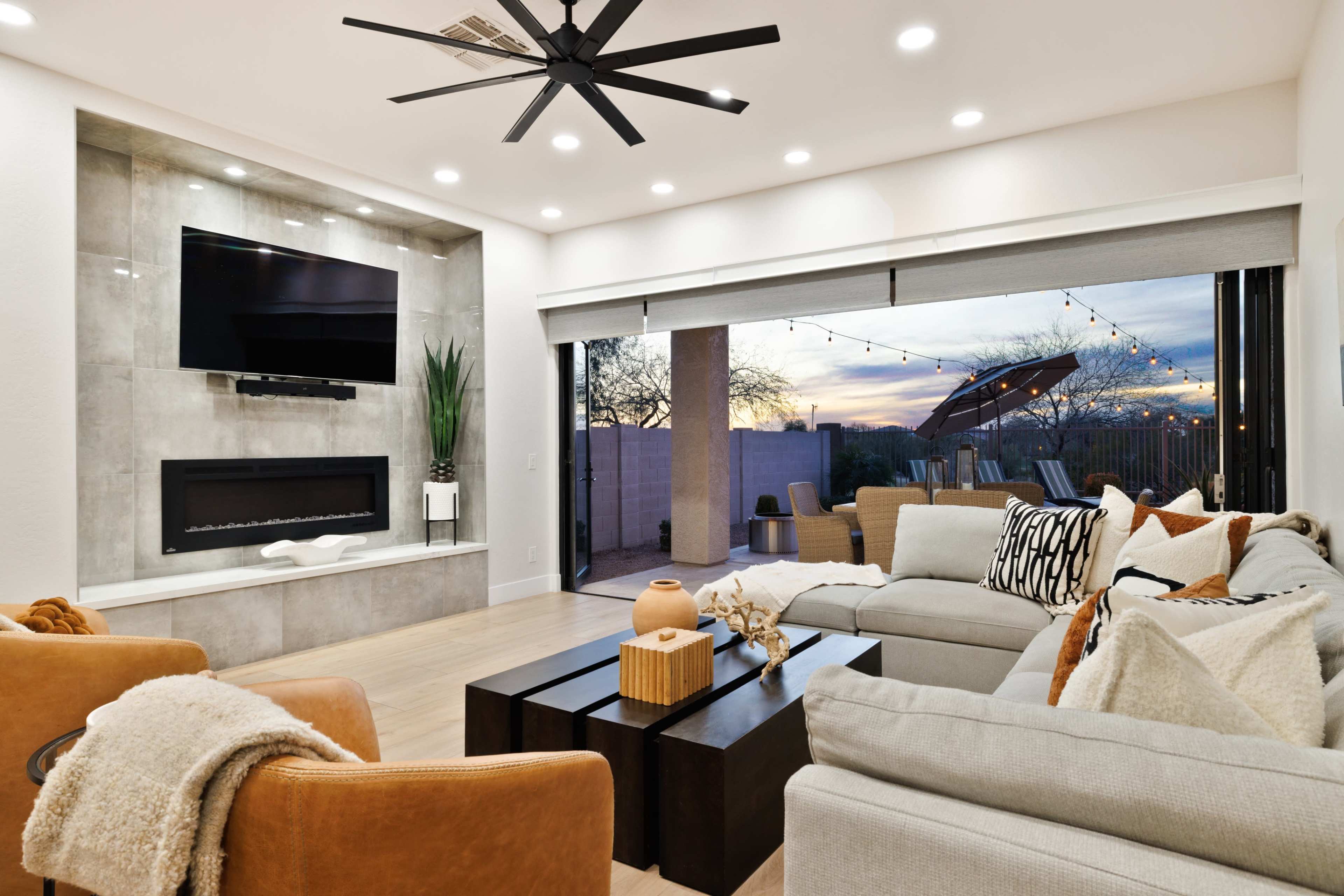 The image shows a modern living room with a large sectional sofa, a dark coffee table, a mounted television, and a wall displaying a sleek electric fireplace, with large windows revealing an outdoor patio at sunset.