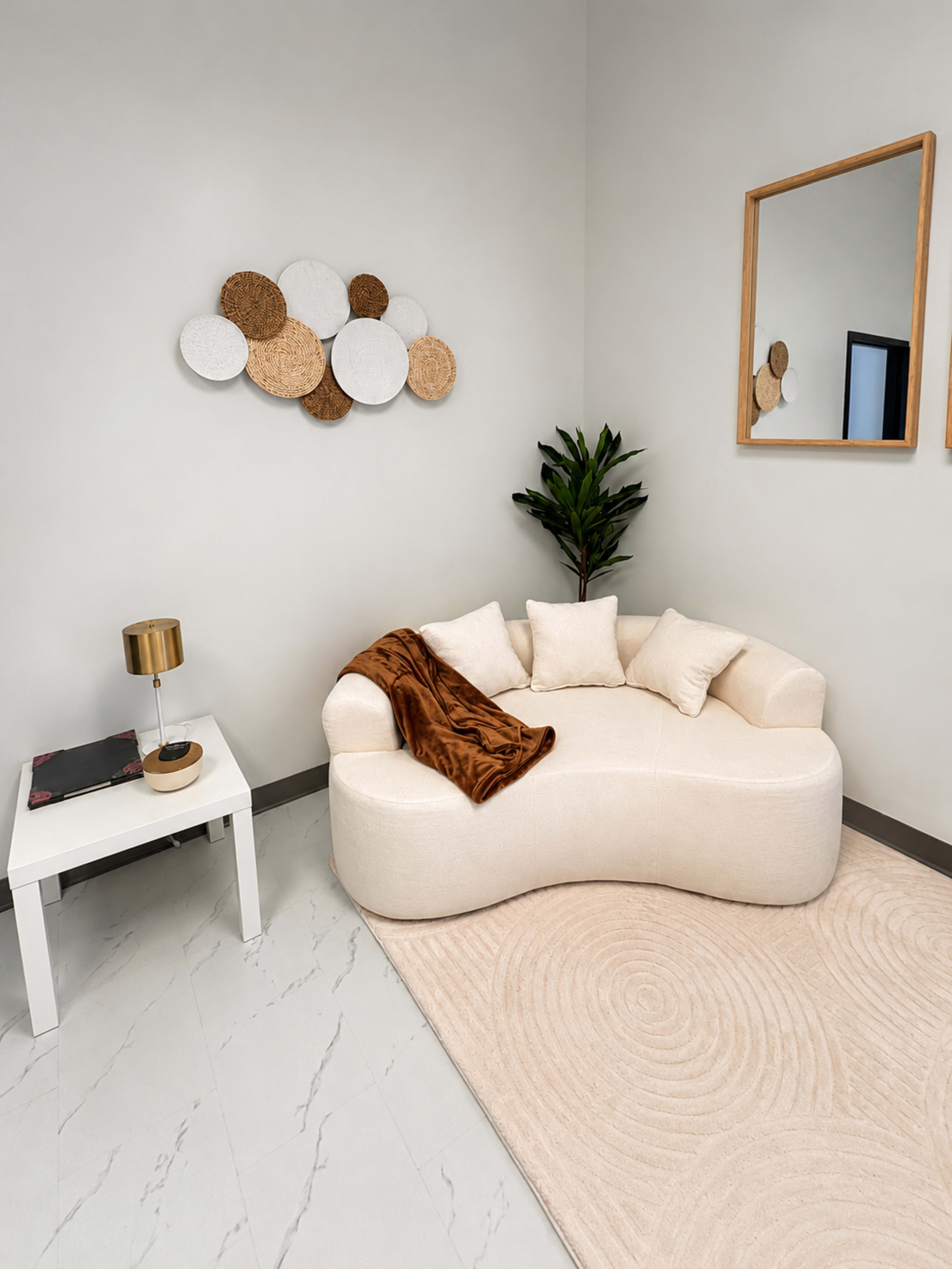 A modern corner seating area features a curved cream-colored couch with decorative pillows, a small white side table, a lamp, and a rug.