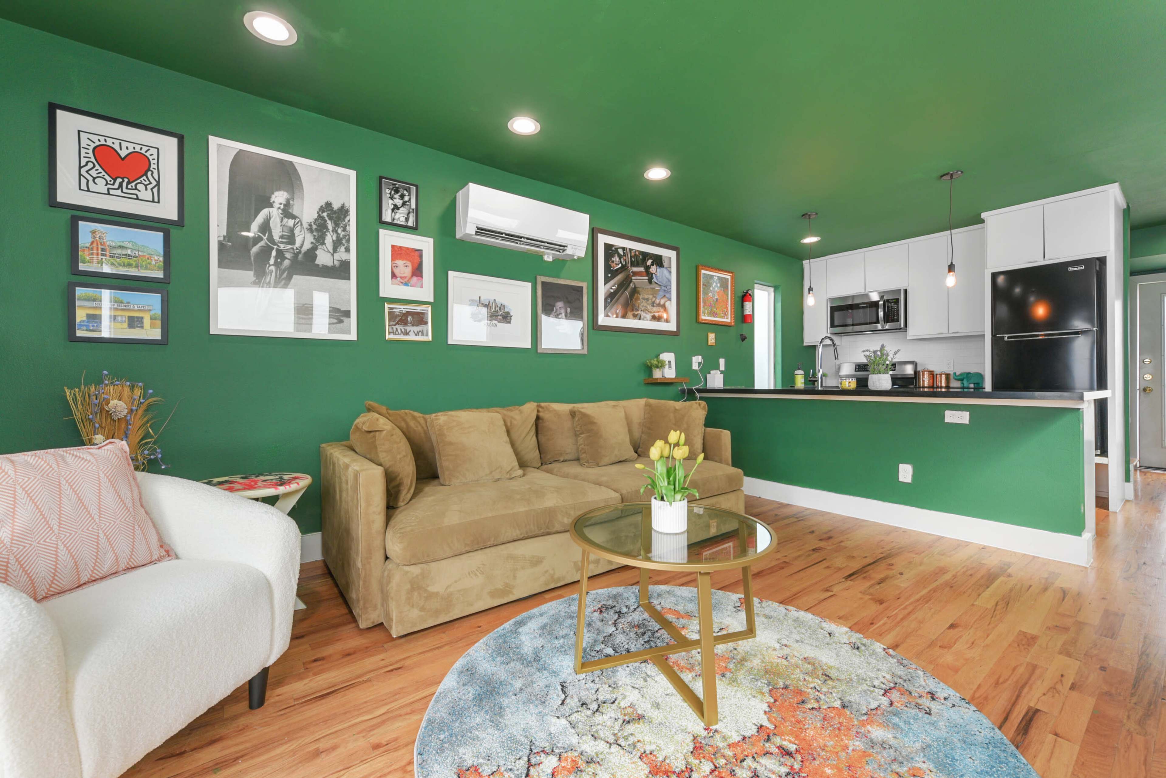 The image shows a cozy living space featuring a green wall, a beige sofa, a round rug with abstract patterns, and a kitchen area with modern appliances and framed artwork on the walls.