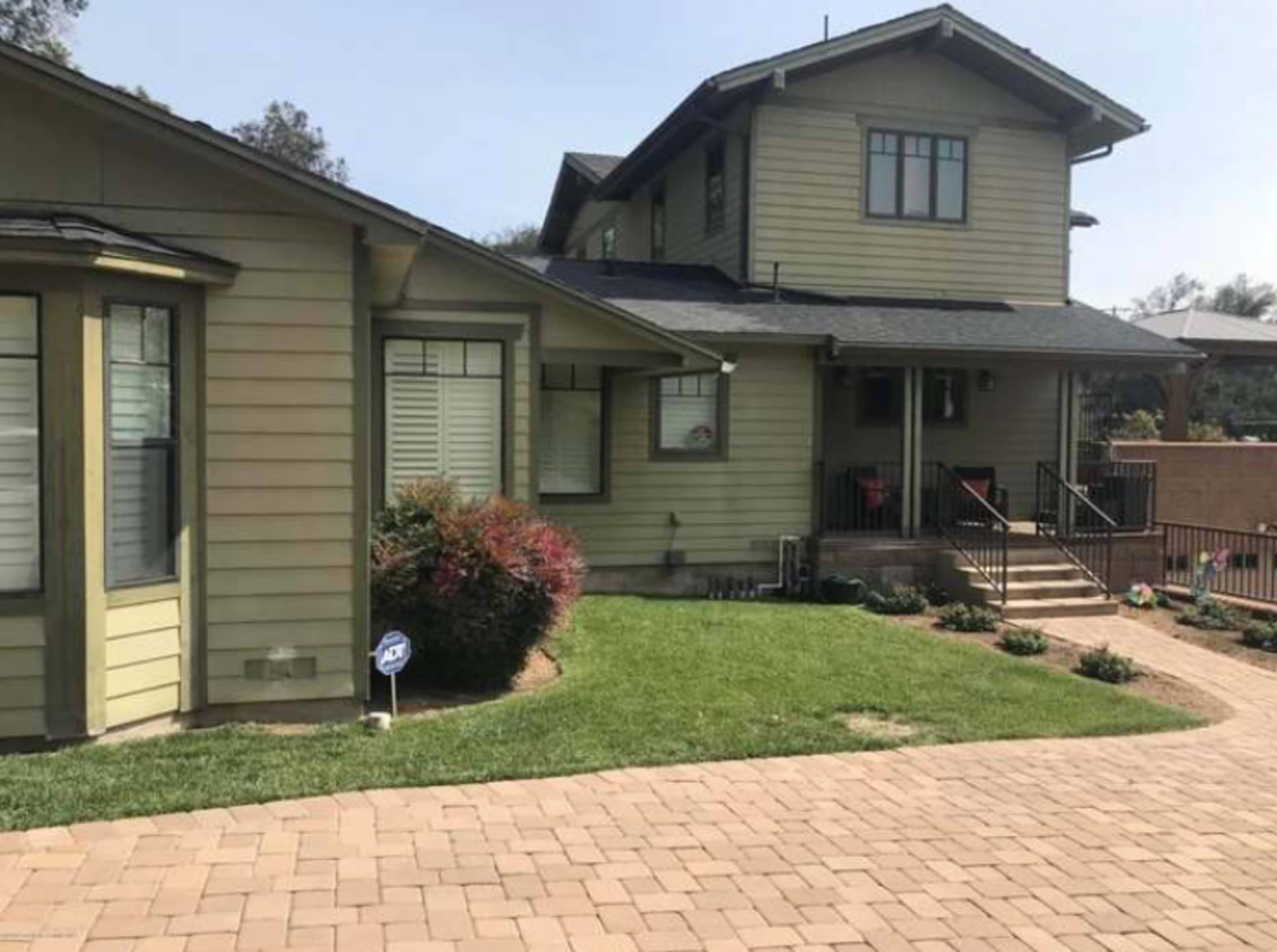 A two-story green house with a porch and landscaped yard, surrounded by a brick pathway.