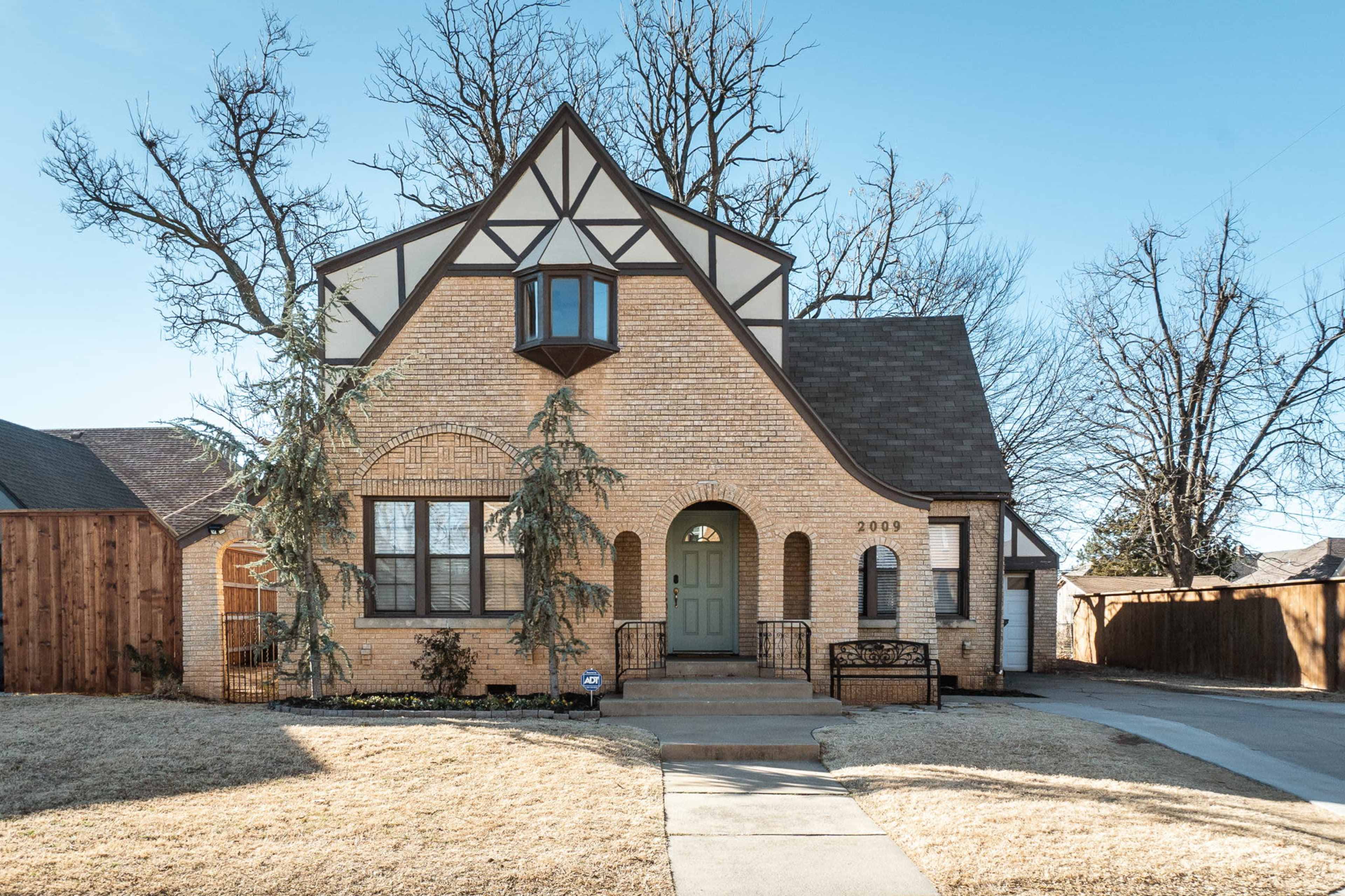 A two-story brick house with a steeply pitched roof, featuring a prominent bay window and symmetrical front yard landscaping.