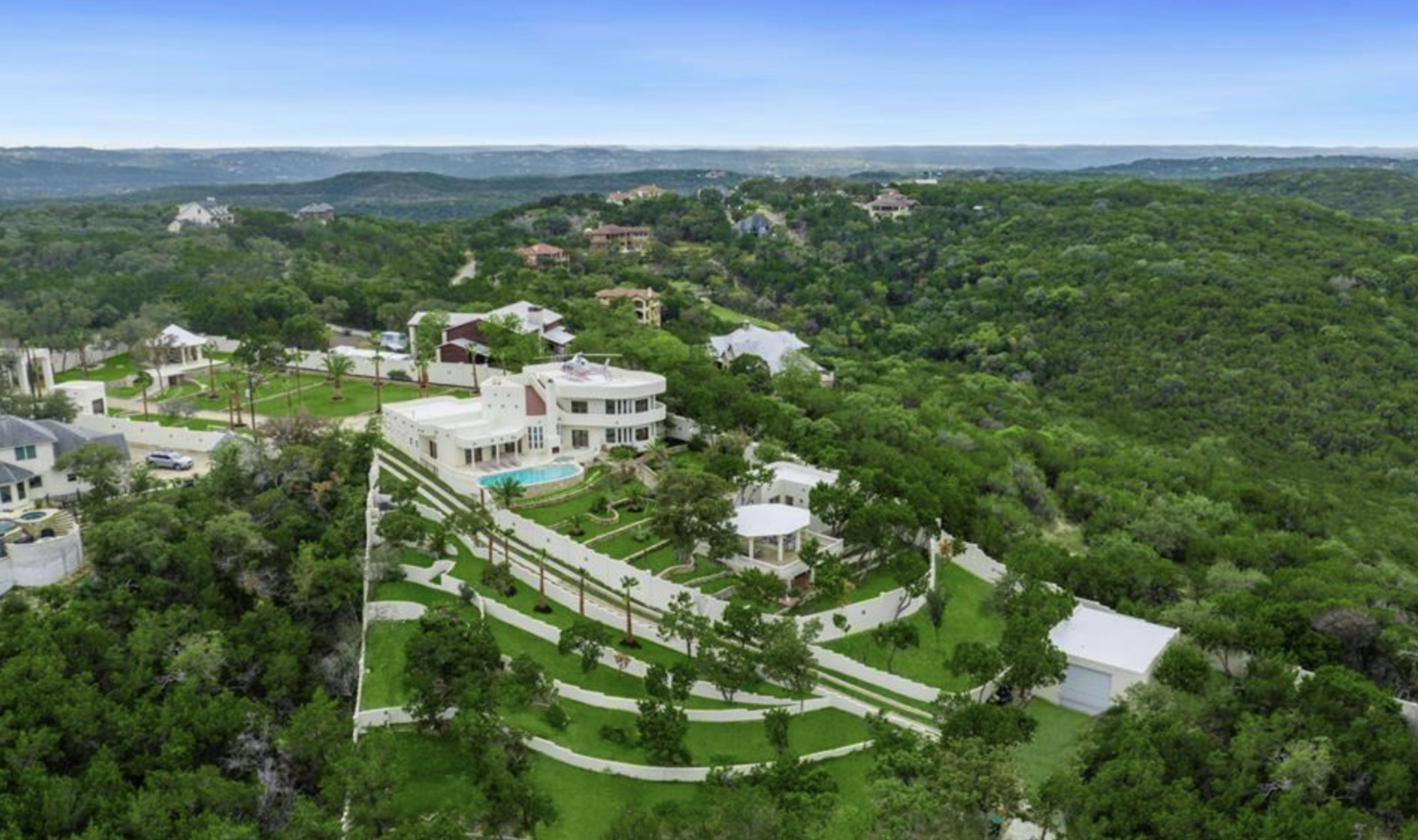 A large modern mansion situated on a hillside surrounded by lush greenery and other homes in the distance.