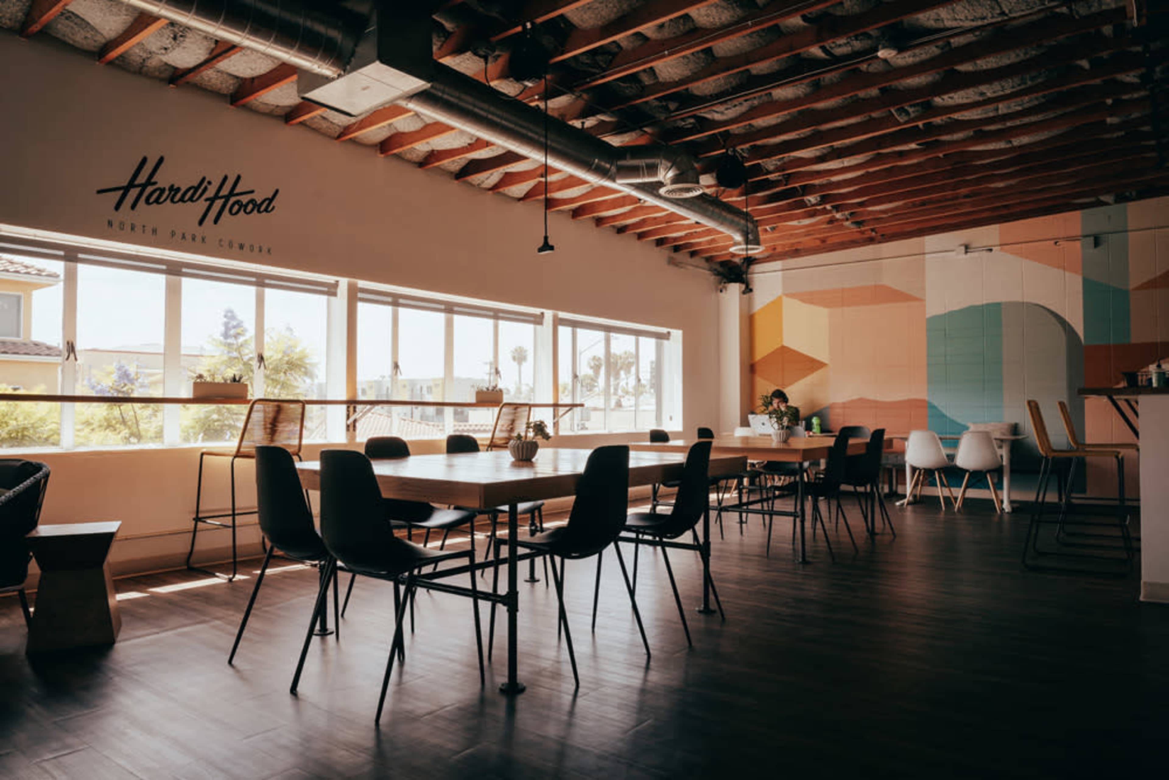A spacious, modern workspace features wooden tables and chairs, large windows allowing natural light, and a colorful wall mural.