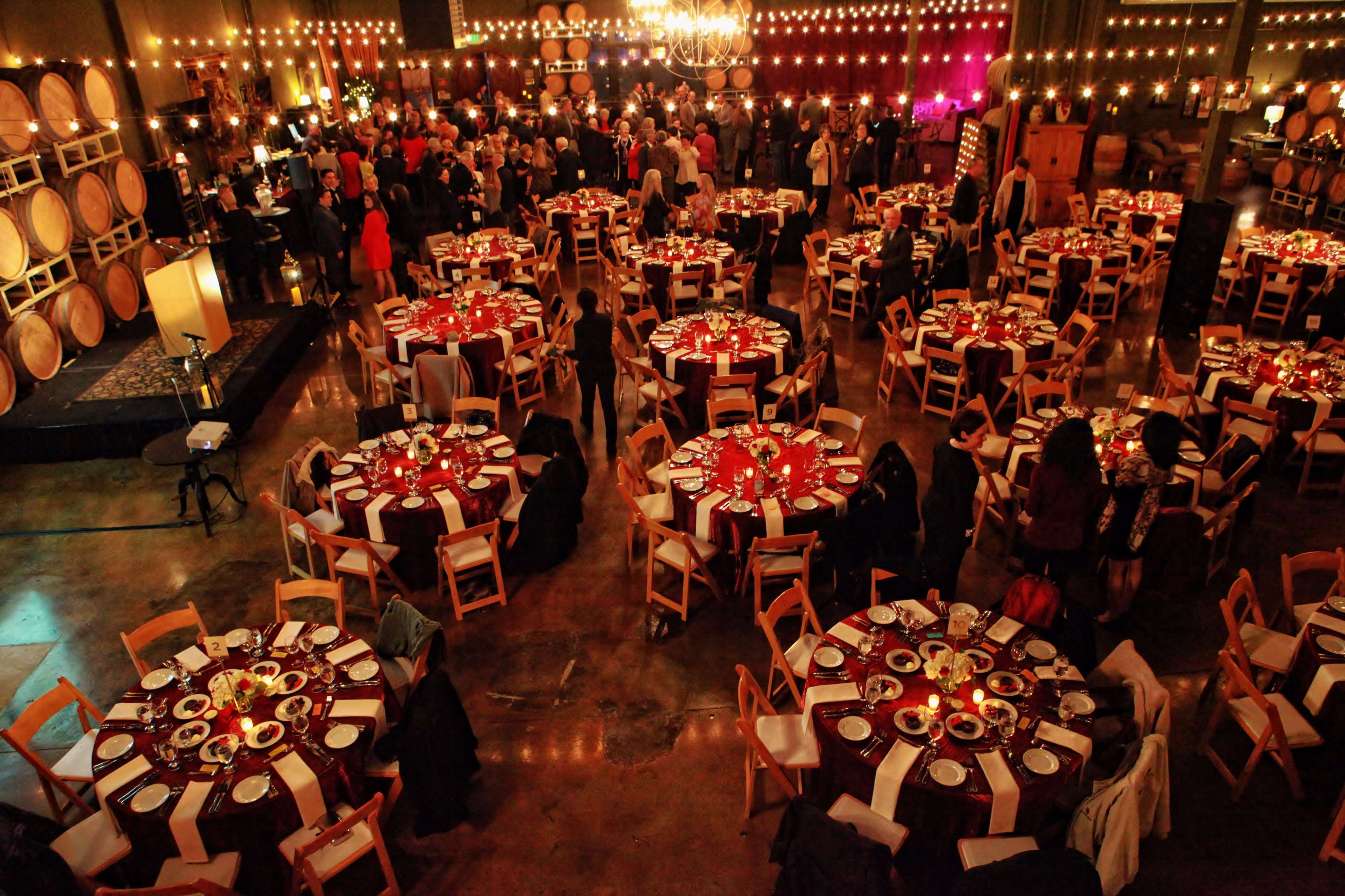A large banquet hall filled with people gathered around elegantly set round tables, with decorations and string lights in the background.
