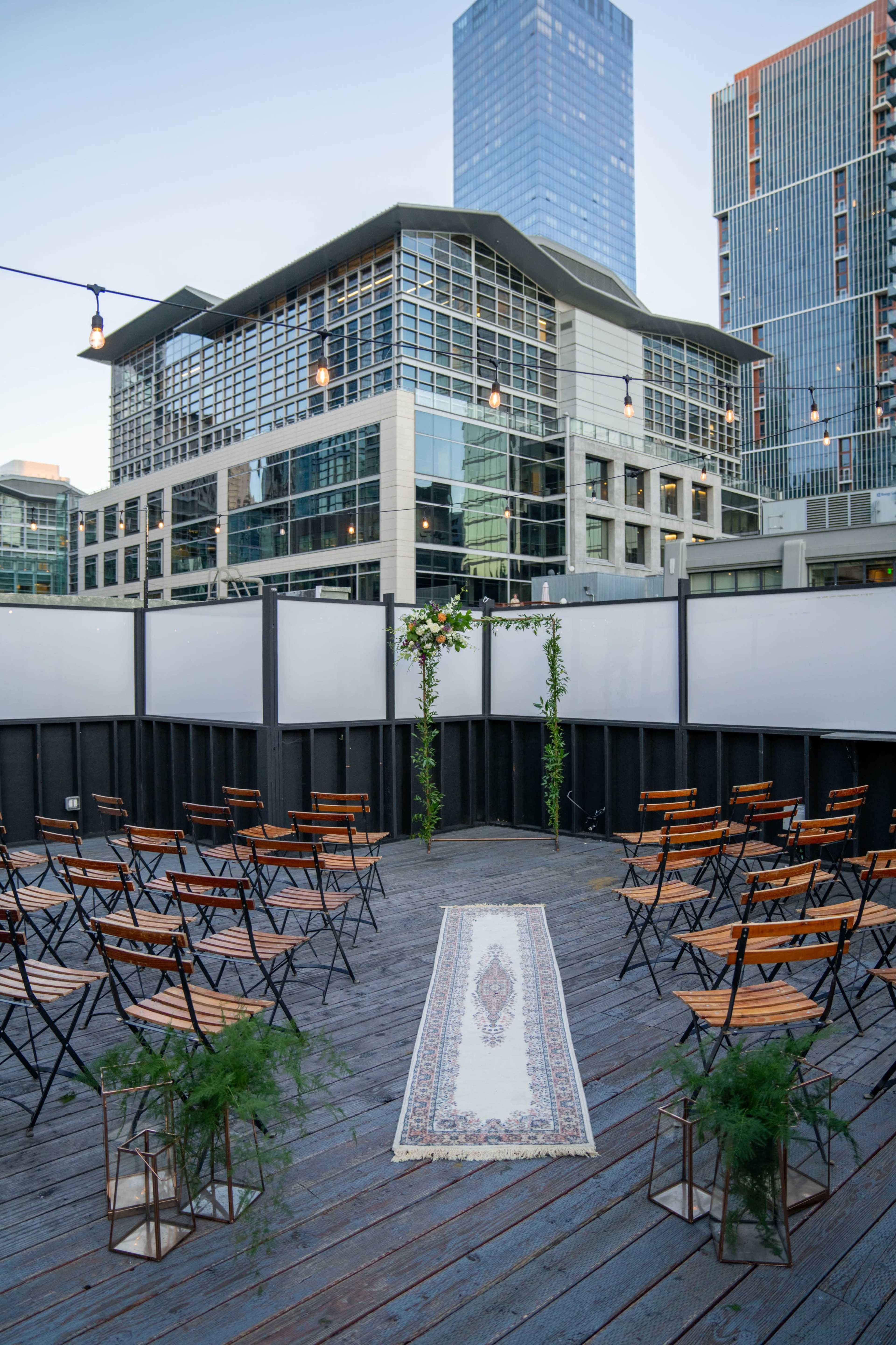 An outdoor event space with wooden chairs arranged in rows facing a decorative arch, set against a backdrop of modern buildings.