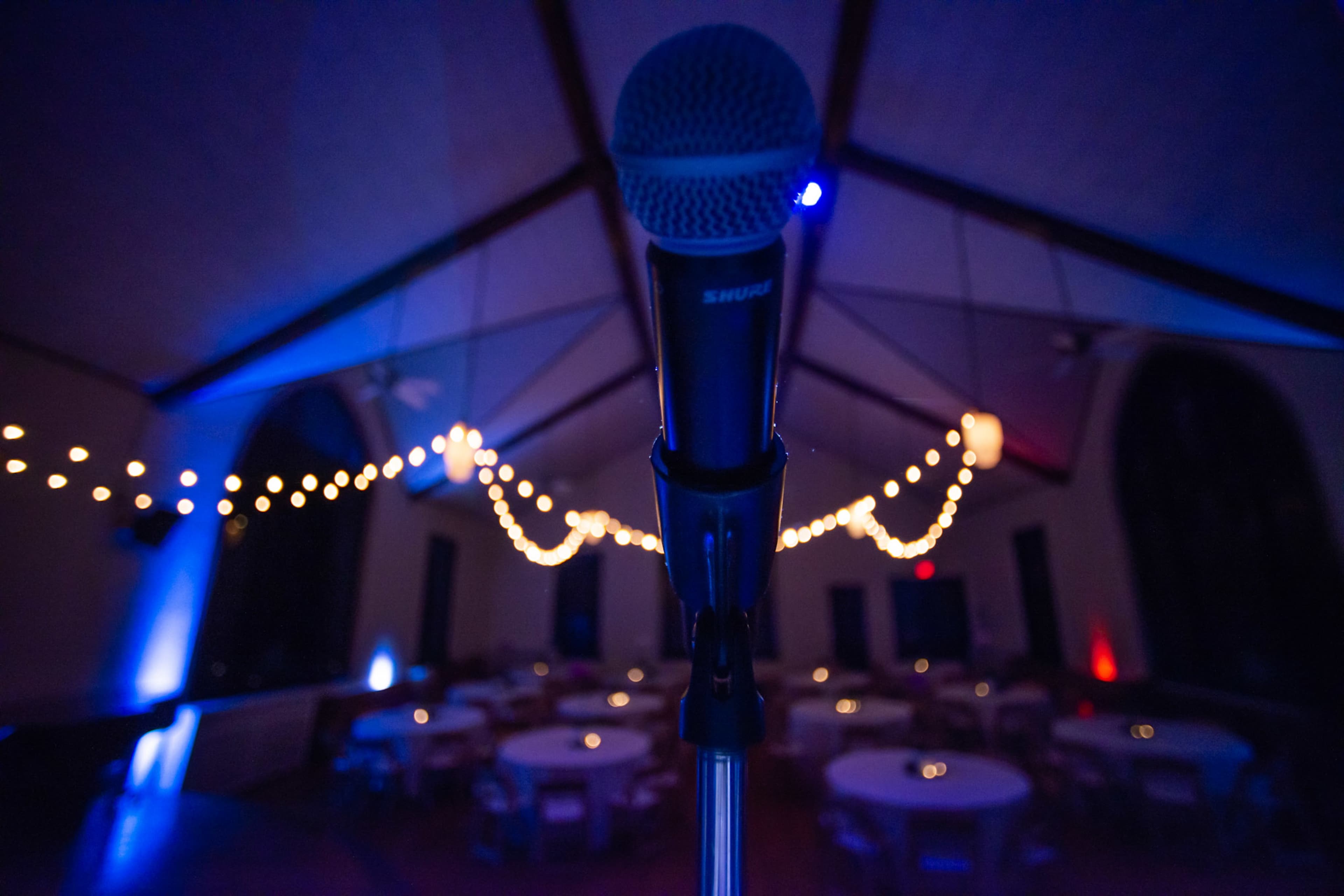 A microphone stands in the foreground with a decorated venue featuring tables and string lights in the background.
