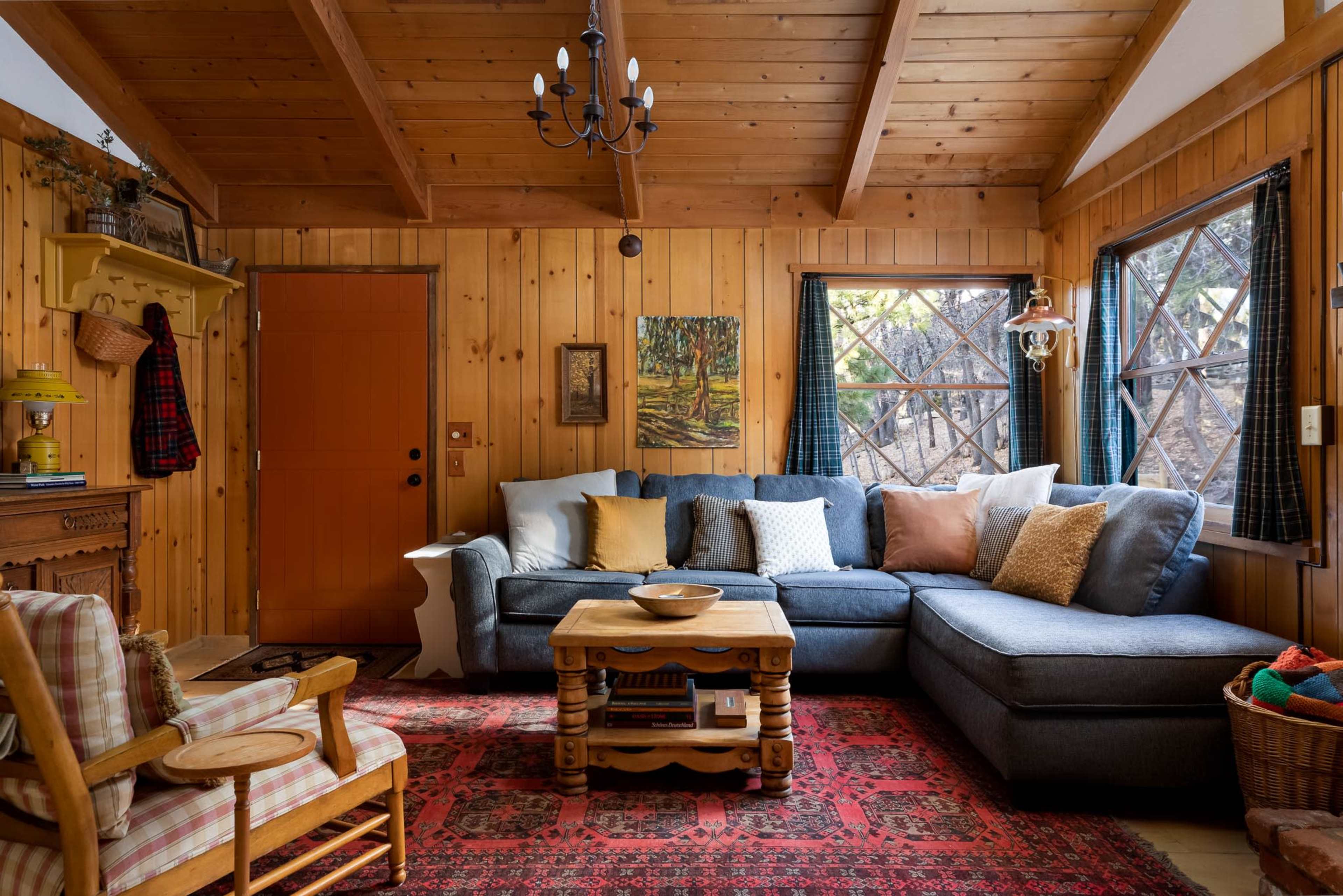 The scene depicts a cozy living room with wooden walls and a sectional couch, a coffee table in the center, and a door leading outside.