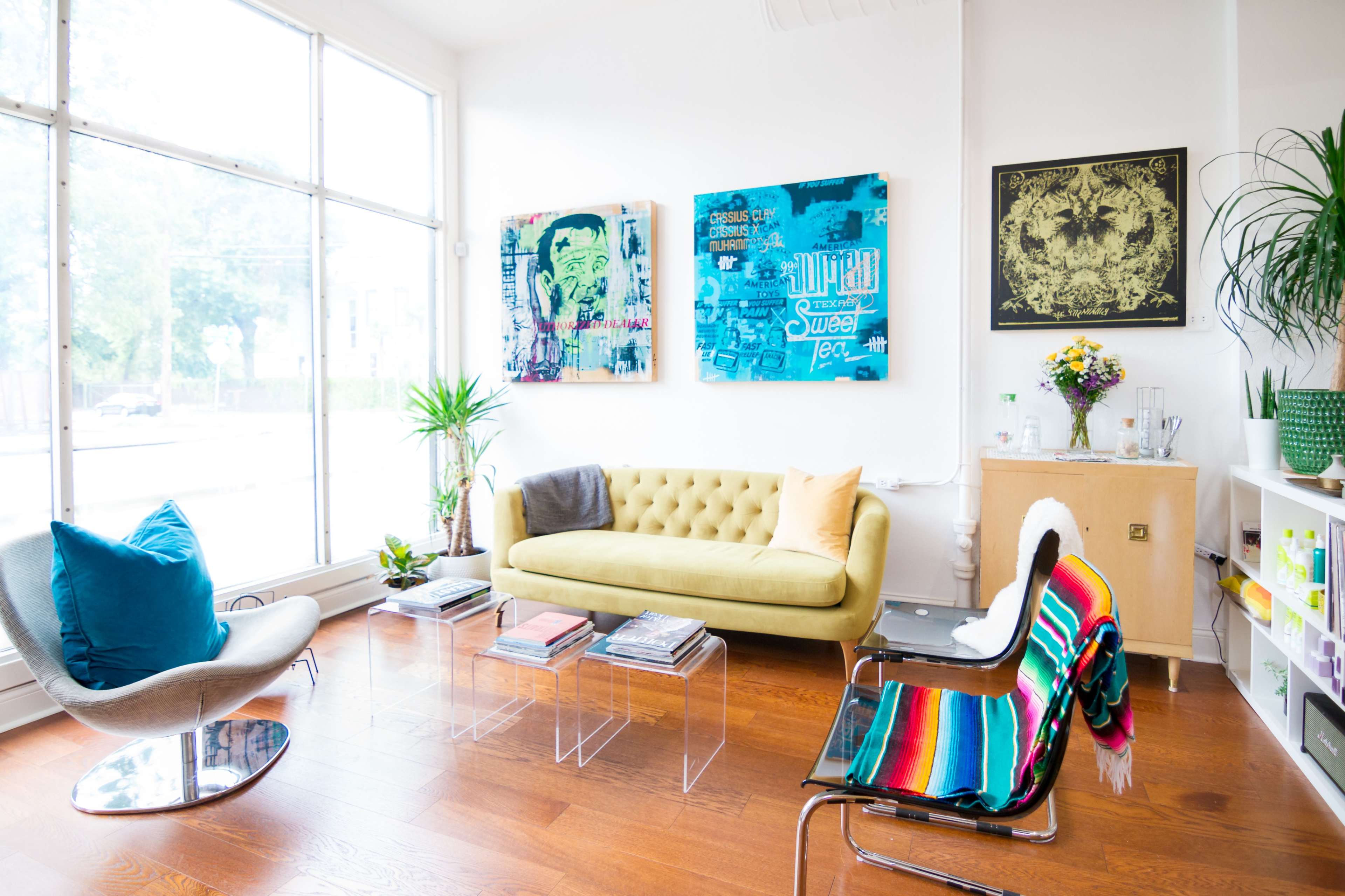 A brightly lit living room with a yellow tufted sofa, colorful artwork on the walls, and a mix of modern furniture, including a clear coffee table and a striped chair.