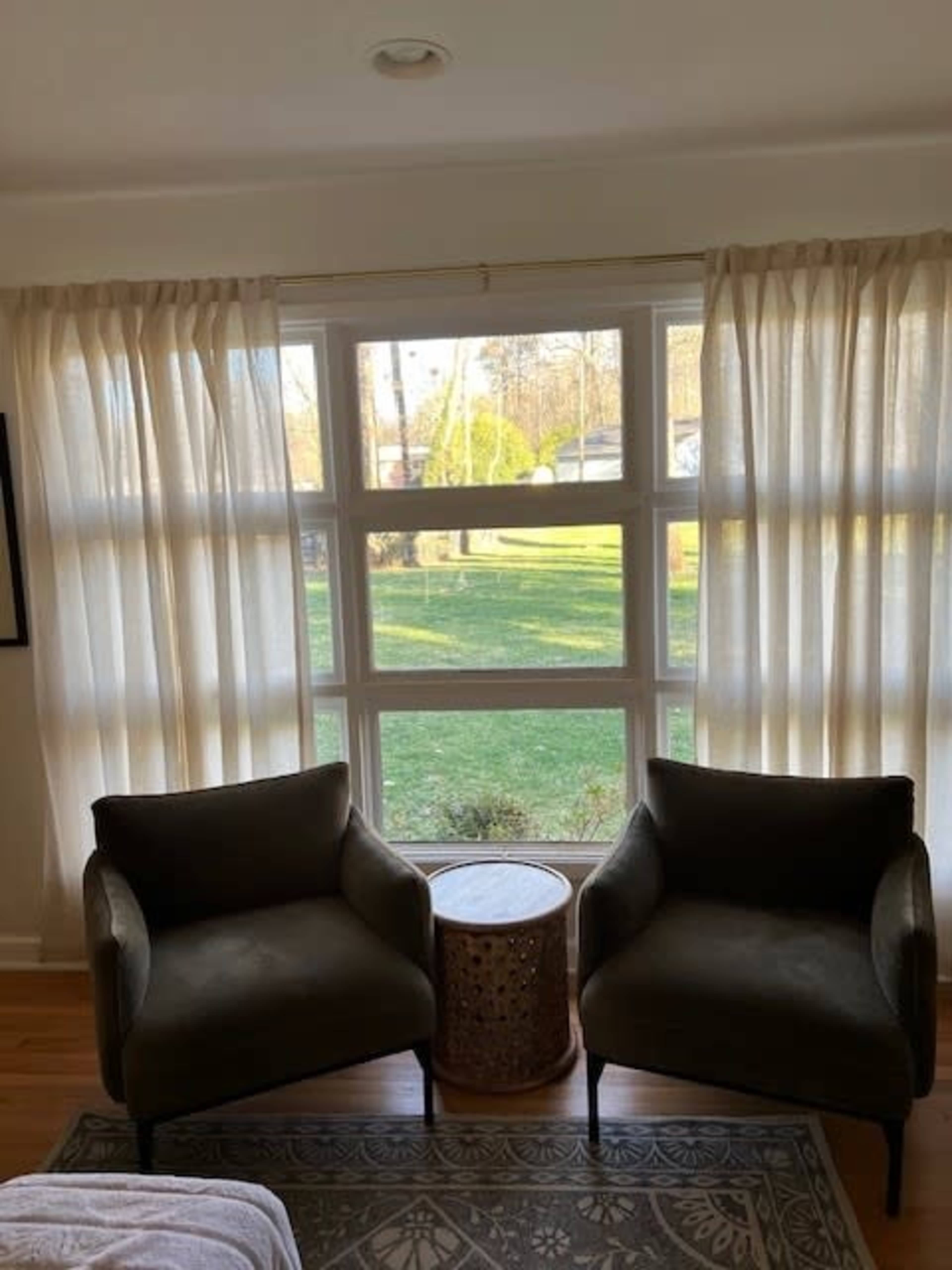 Two green armchairs are positioned near a large window with sheer curtains, revealing a view of a grassy yard outside.