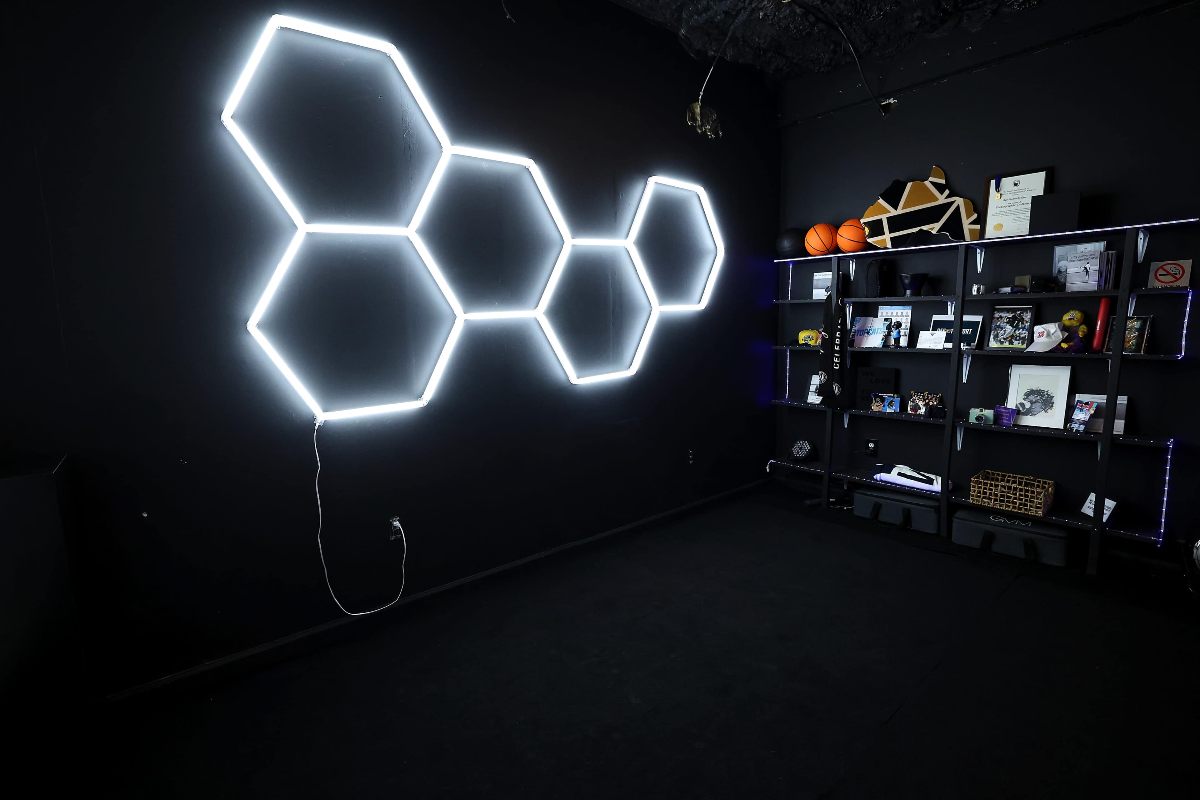 The image shows a black room featuring a hexagonal neon light installation on one wall and a black shelving unit displaying various items, including sports equipment and framed documents.