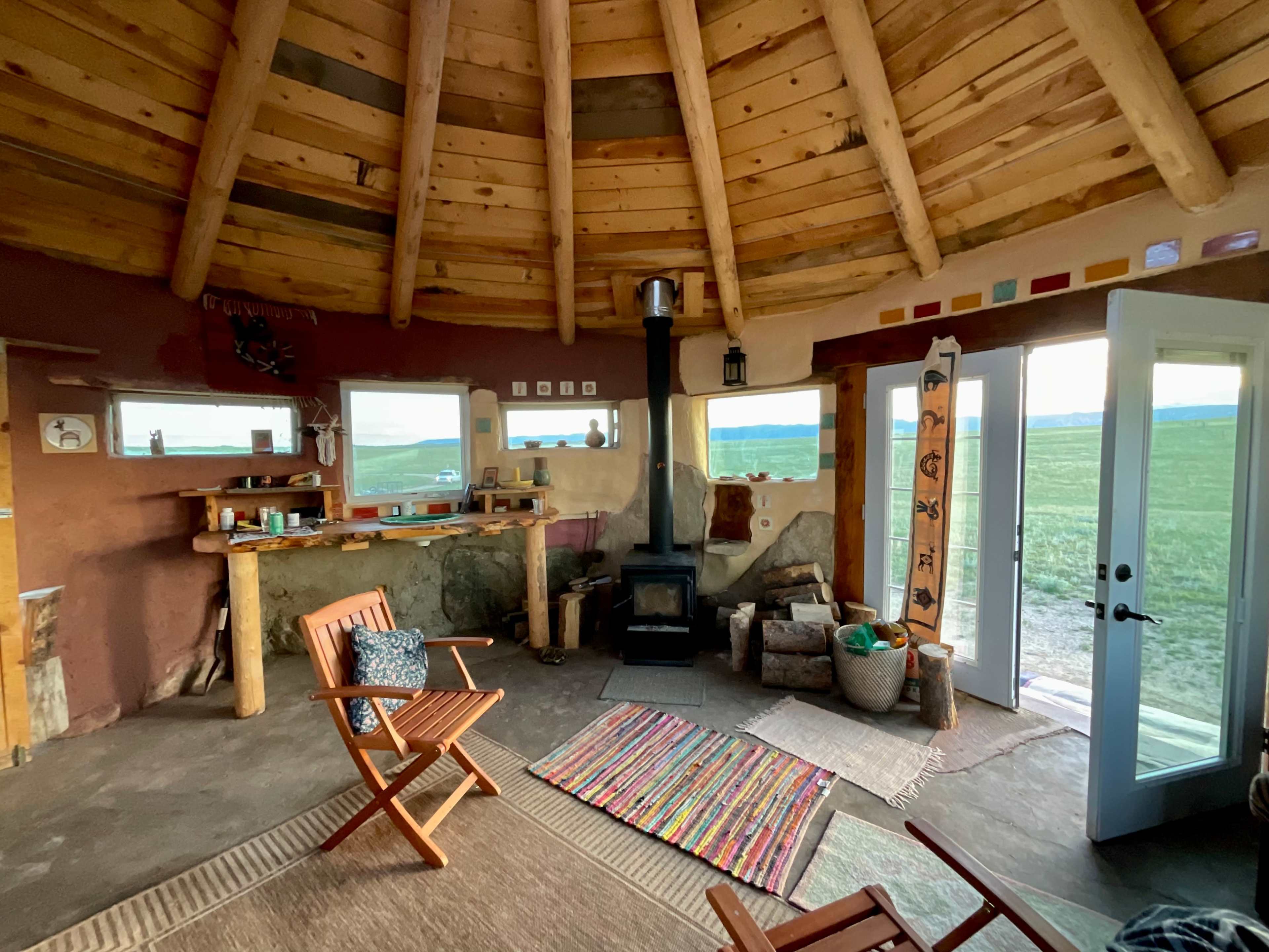The interior of a circular, earth-sheltered dwelling features a central wood stove, wooden chairs, and large windows that overlook a grassy landscape.