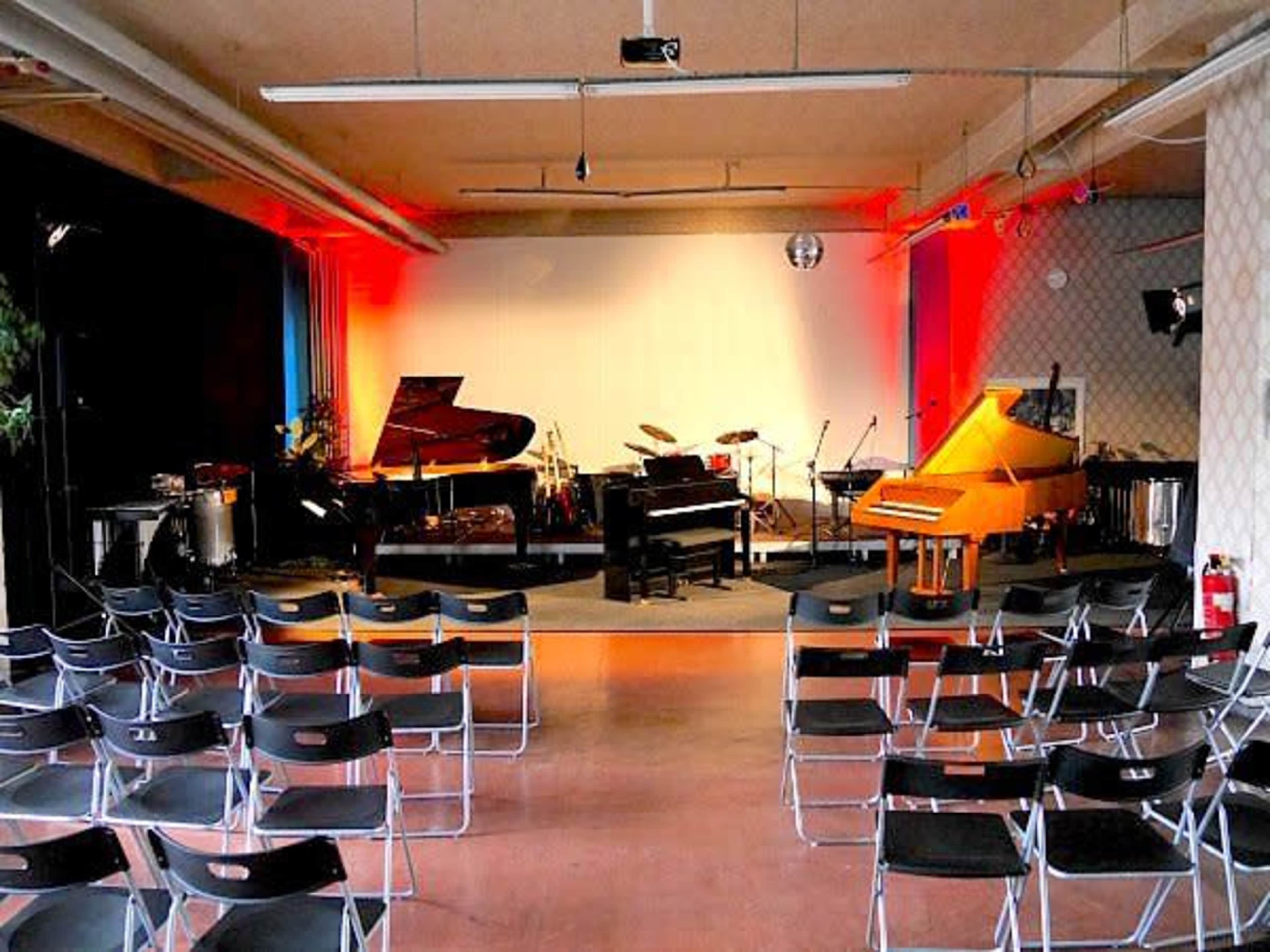 The image shows a music performance space with several pianos and a set of empty black chairs arranged in rows facing the stage.