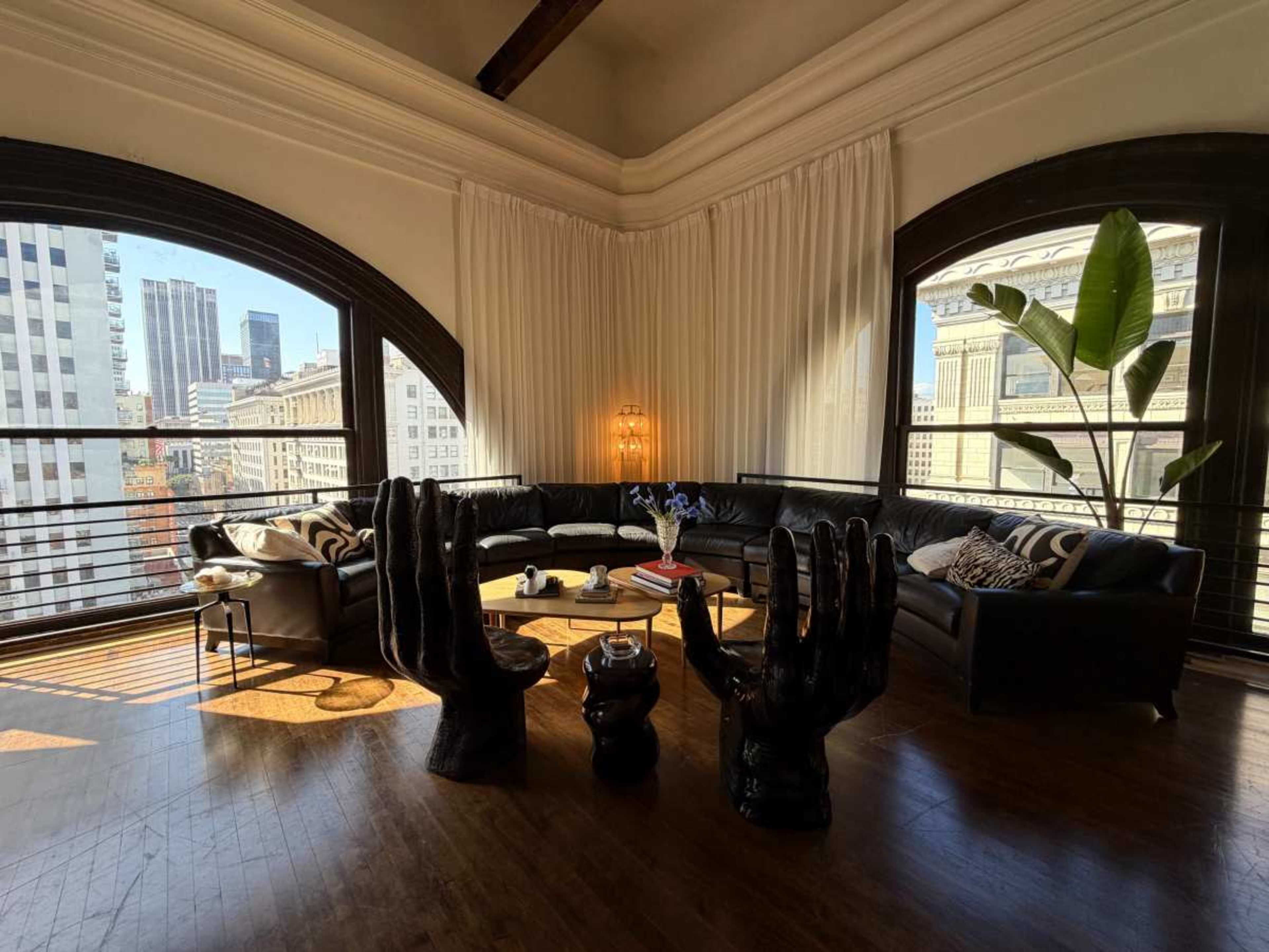 A modern living room features a curved sectional sofa, unique hand-shaped chairs, and large windows with views of a city skyline.