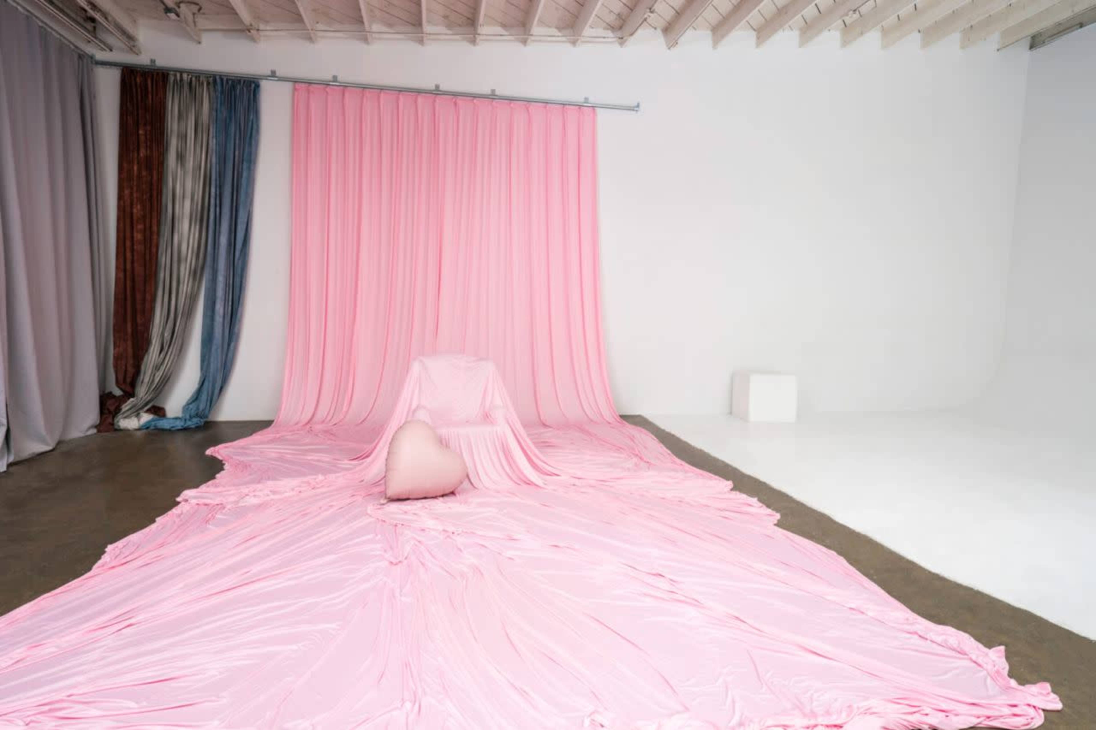 The image shows a spacious studio with a pink fabric draping from the ceiling and covering the floor, complemented by a single heart-shaped prop.
