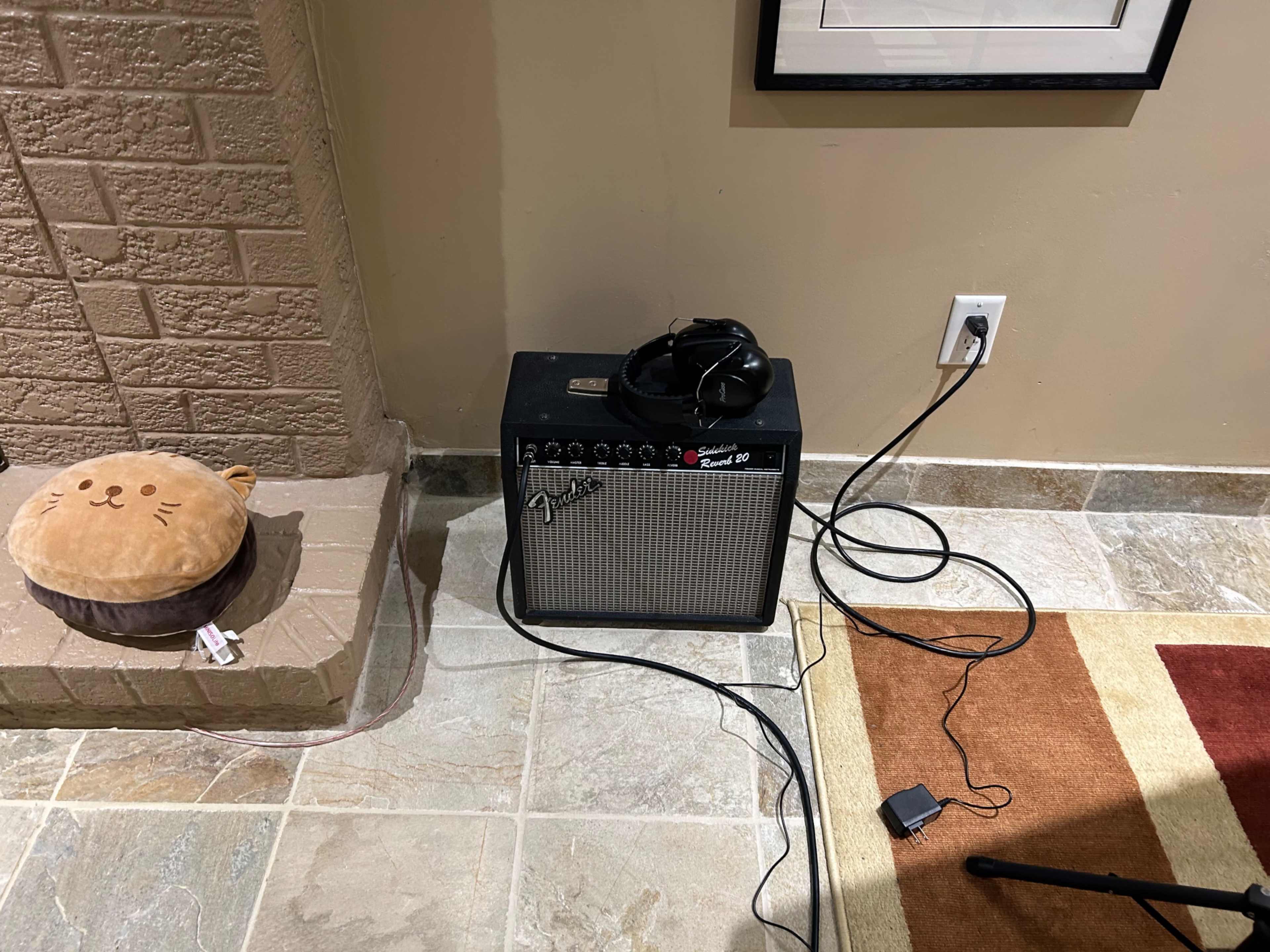 A guitar amplifier with headphones resting on top, placed next to a plush cat-shaped cushion and surrounded by cables on a tiled floor.