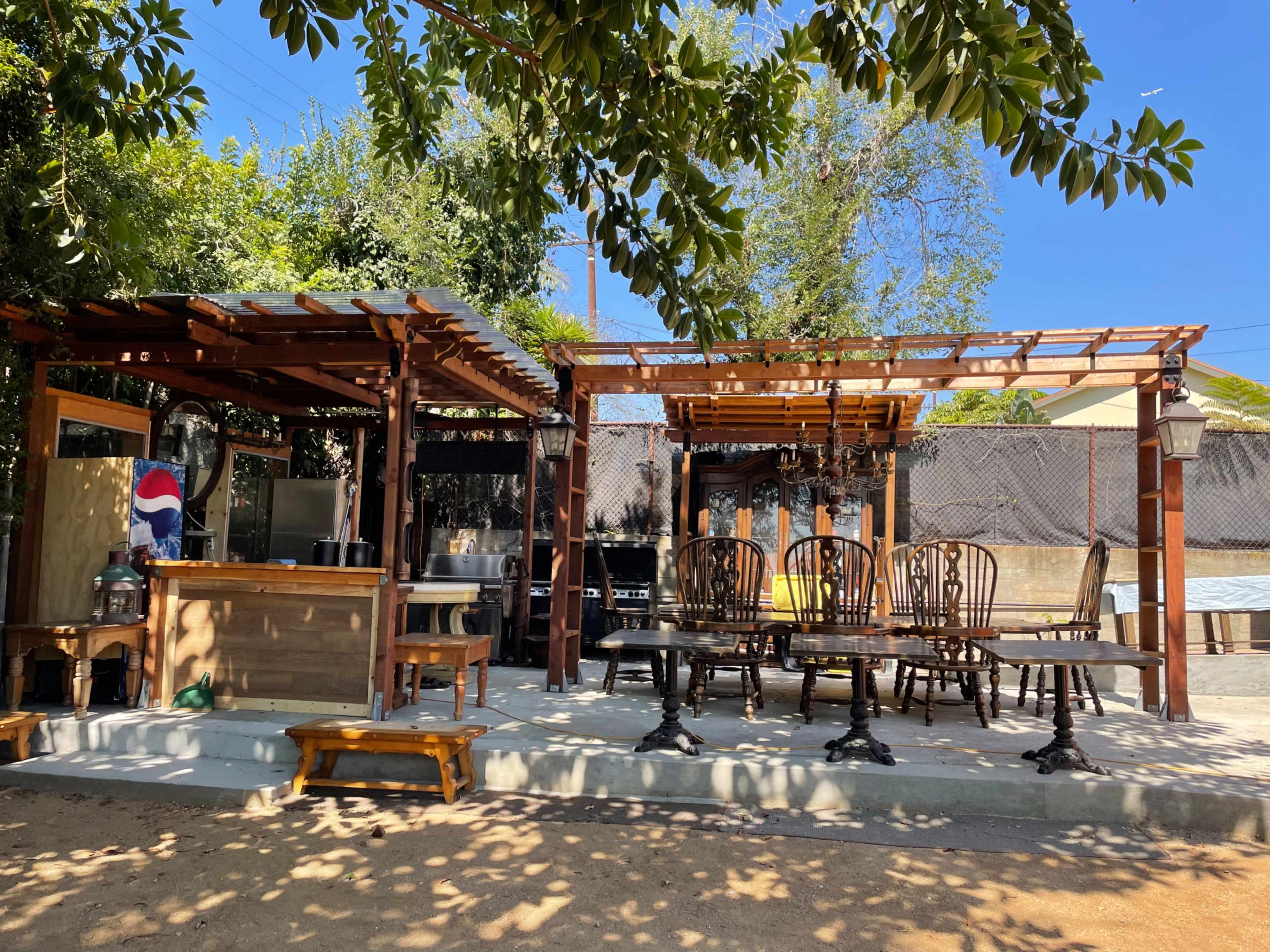 The image shows an outdoor seating area with wooden furniture and a bar structure, surrounded by trees and sunlight.
